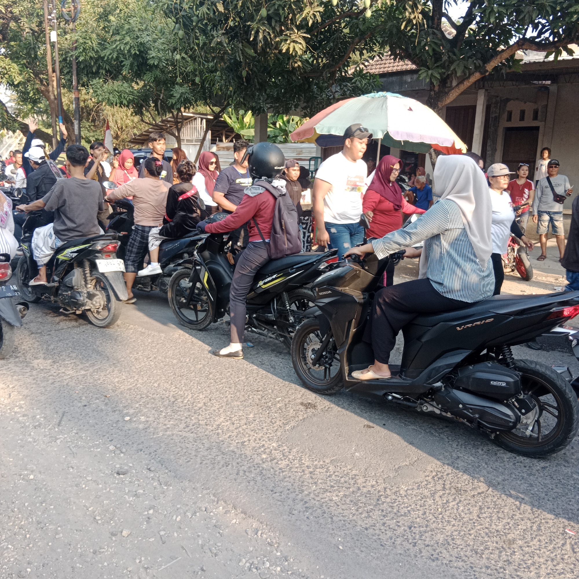 In the city of Tegalrejo, Indonesia, a street scene unfolds with a crowd of people and motorcycles. The environment appears to be outdoors, with sunlight casting shadows on the asphalt road. Trees with green foliage are visible in the background, suggesting a warm climate. Buildings with visible roofing and walls line the street.
The primary subjects are individuals, many of whom are riding or standing near motorcycles. The people are dressed in casual attire, with a mix of light and dark clothing. Some are wearing headscarves, common in Indonesia. A few individuals are clearly depicted as drivers or passengers on the motorcycles, while others are observing or interacting within the group. The motorcycles themselves are varied, with a black Honda scooter being a prominent feature in the foreground.
The action appears to be a gathering or a momentary stop, possibly due to traffic or a social interaction. People are looking in various directions, and some seem to be engaged in conversation. The presence of motorcycles suggests transportation and daily life activities in this urban or semi-urban setting. The overall impression is a snapshot of a community in motion, with a focus on the everyday use of motorcycles as a mode of transport.