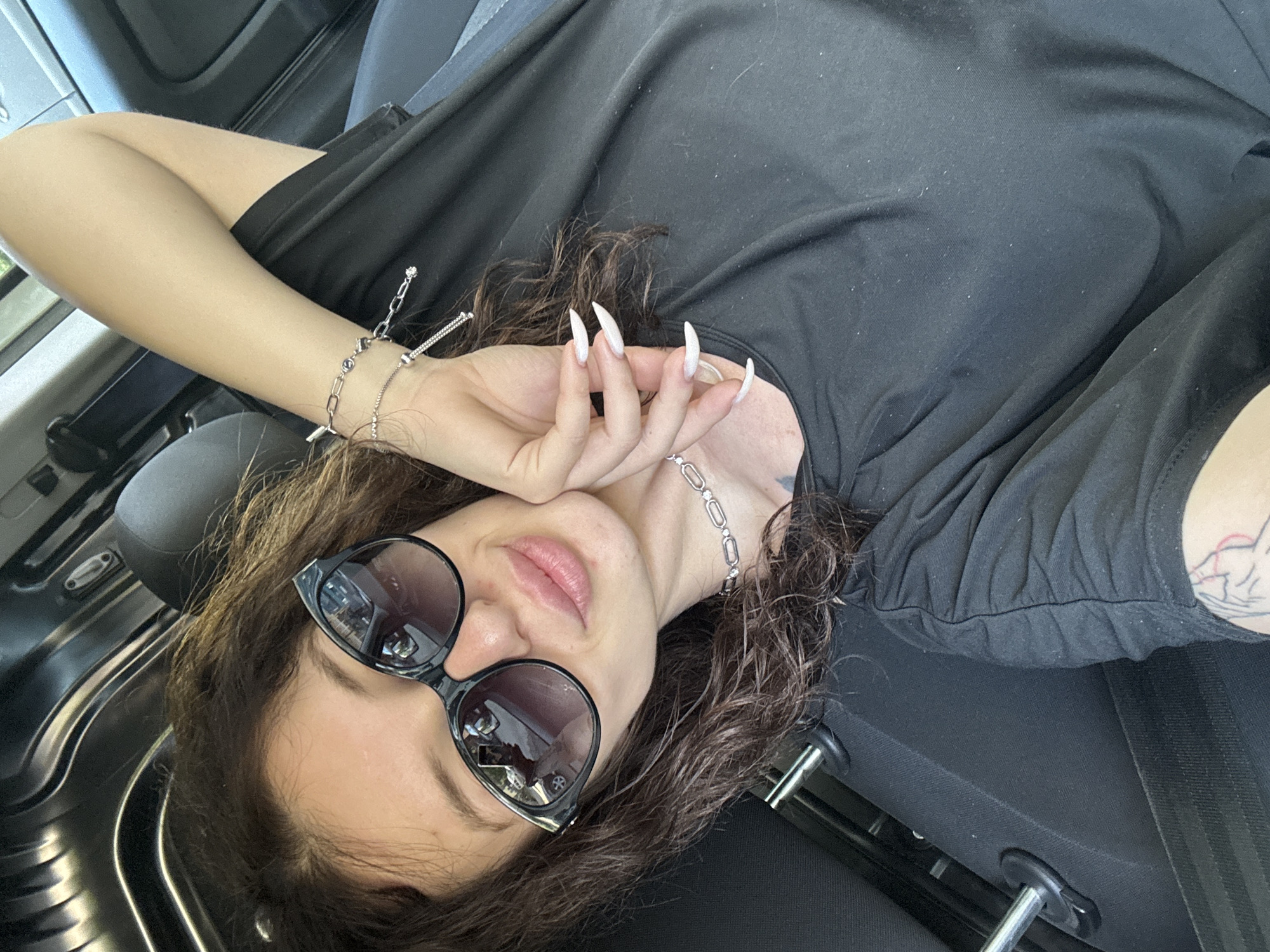 A woman with brown, curly hair and dark sunglasses is lying down in the passenger seat of a vehicle. She is wearing a black t-shirt and a silver chain necklace. Her fingernails are long and white. She is wearing a silver bracelet on her left wrist. The interior of the vehicle is dark gray. A portion of the car door and safety belt are visible. The location is Unknown, Unknown.