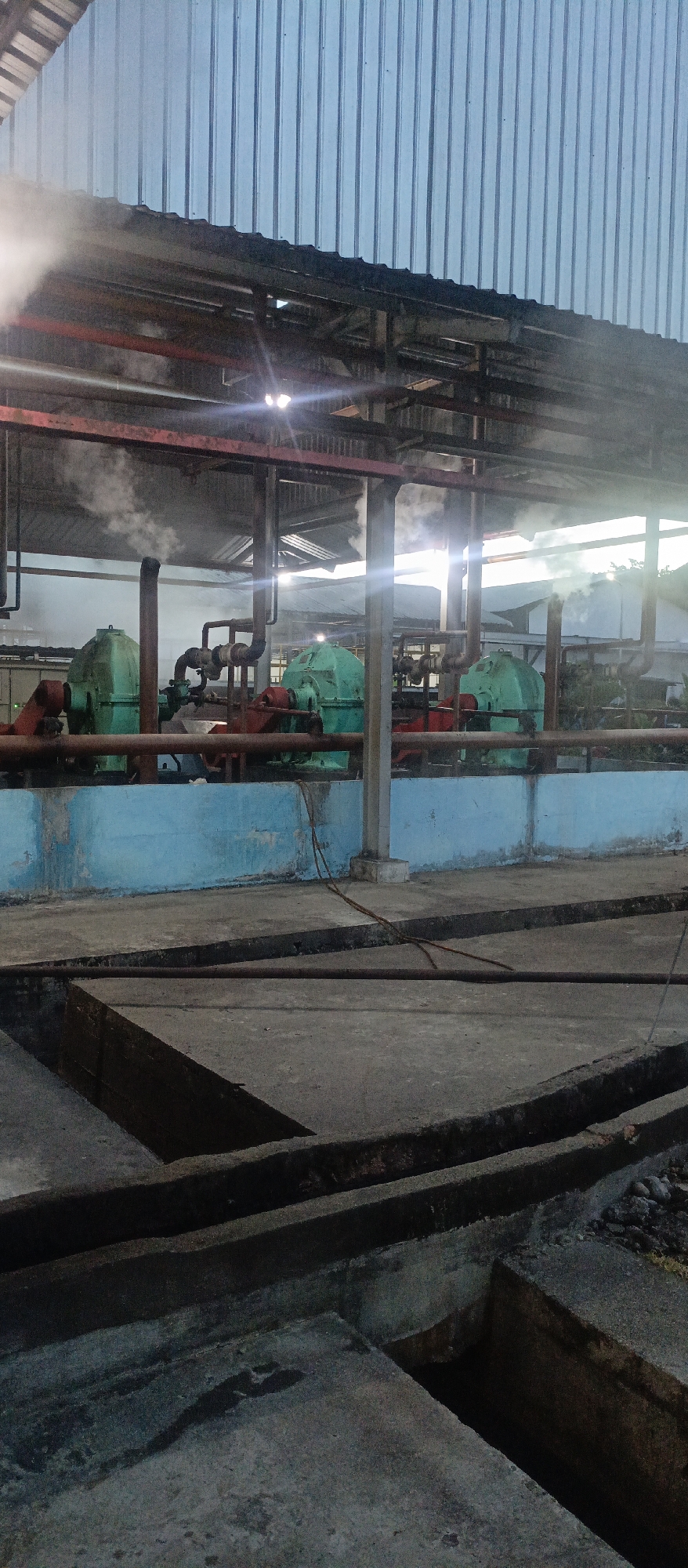 The image depicts an industrial setting, likely a processing plant, with heavy machinery and piping visible. Three large, green machines, possibly pumps or generators, are positioned along a light blue wall. Red pipes and supports are prominent throughout the scene, and steam or smoke is emanating from several points, suggesting active operations. The ground is concrete, with visible channels or trenches. The overhead structure has a corrugated metal roof and exposed beams. The lighting suggests either early morning or late evening. This scene is located in the city of Iemirah, Indonesia.