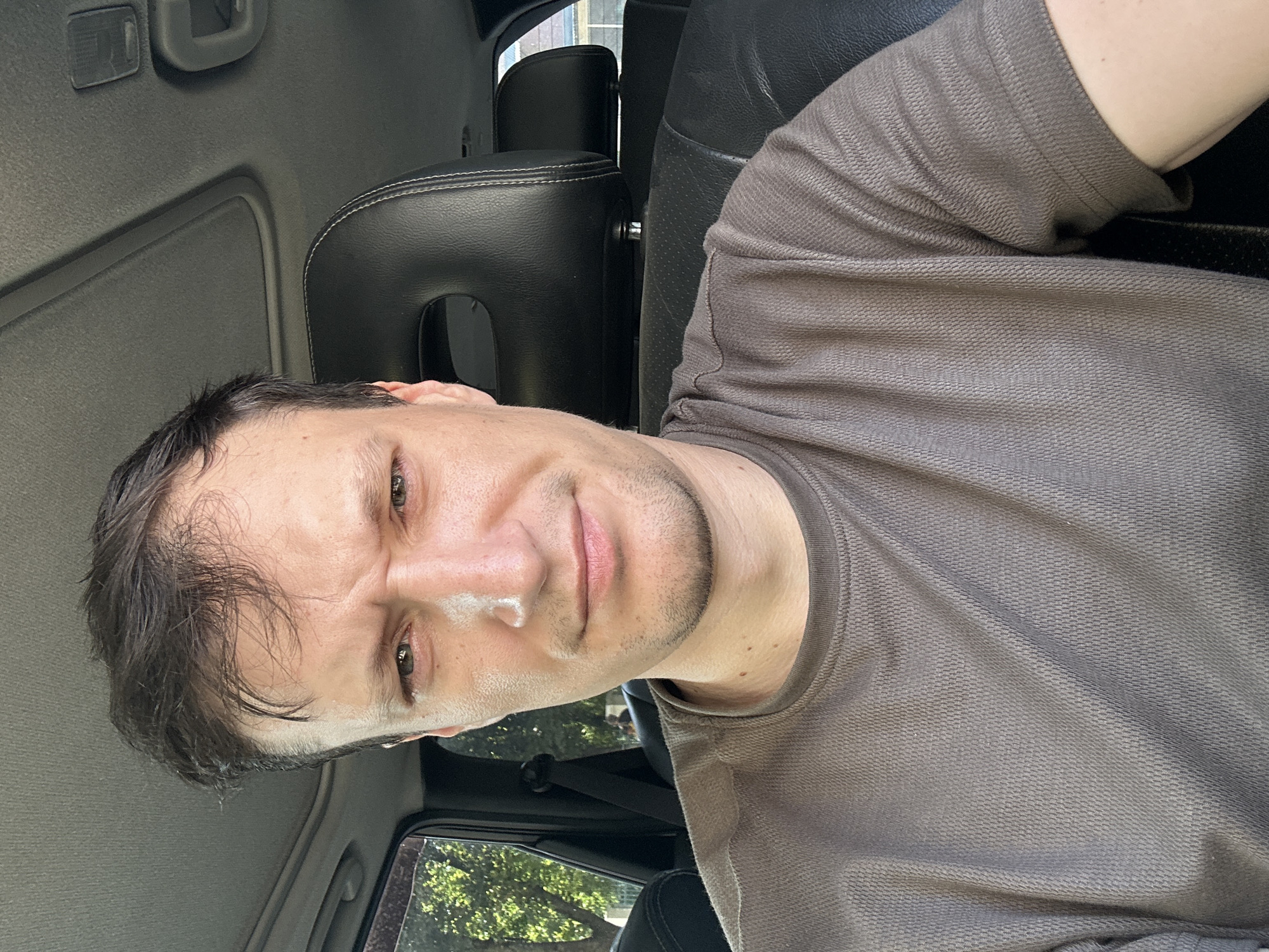 A medium shot captures a man from the chest up, seated inside a car. He has dark, short hair with a receding hairline and light skin. His eyebrows are furrowed, and his eyes are open and looking directly at the camera. He has a light stubble on his chin and jawline. He is wearing a brown t-shirt with a textured pattern. The interior of the car is visible, with a black leather headrest behind him and a sunroof in the ceiling. The background outside the car window shows green trees and a blurred street scene, suggesting an urban or suburban environment. It is daytime, with natural light illuminating the scene. The location is identified as Kamianets-Podilskyi, Ukraine.