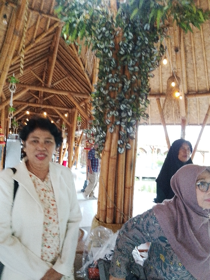 The scene is set within a substantial, open-sided bamboo structure in Pabelan, Indonesia, featuring exposed bamboo beams, trusses, and ceiling panels. In the immediate foreground, a woman with dark, short hair, wearing a white blazer over a light-colored floral blouse, stands facing the camera with a slight smile. To her right and slightly behind, a second woman with glasses wears a light purple hijab and a patterned long-sleeved top, looking towards the right of the frame. Further back and to the right, a third woman wears a black hijab, her face partially obscured, looking away from the camera. A prominent bamboo column, decorated with an artificial leafy vine extending upwards, stands between the foreground woman and the background figures. Beyond this column, a man in a plaid shirt and light trousers is visible walking away from the camera. The background reveals an expanse of water, likely a pond or river, and distant structures. Overhead, exposed incandescent bulbs hang from the bamboo ceiling. Items wrapped in clear plastic are visible on a surface in the lower right foreground.