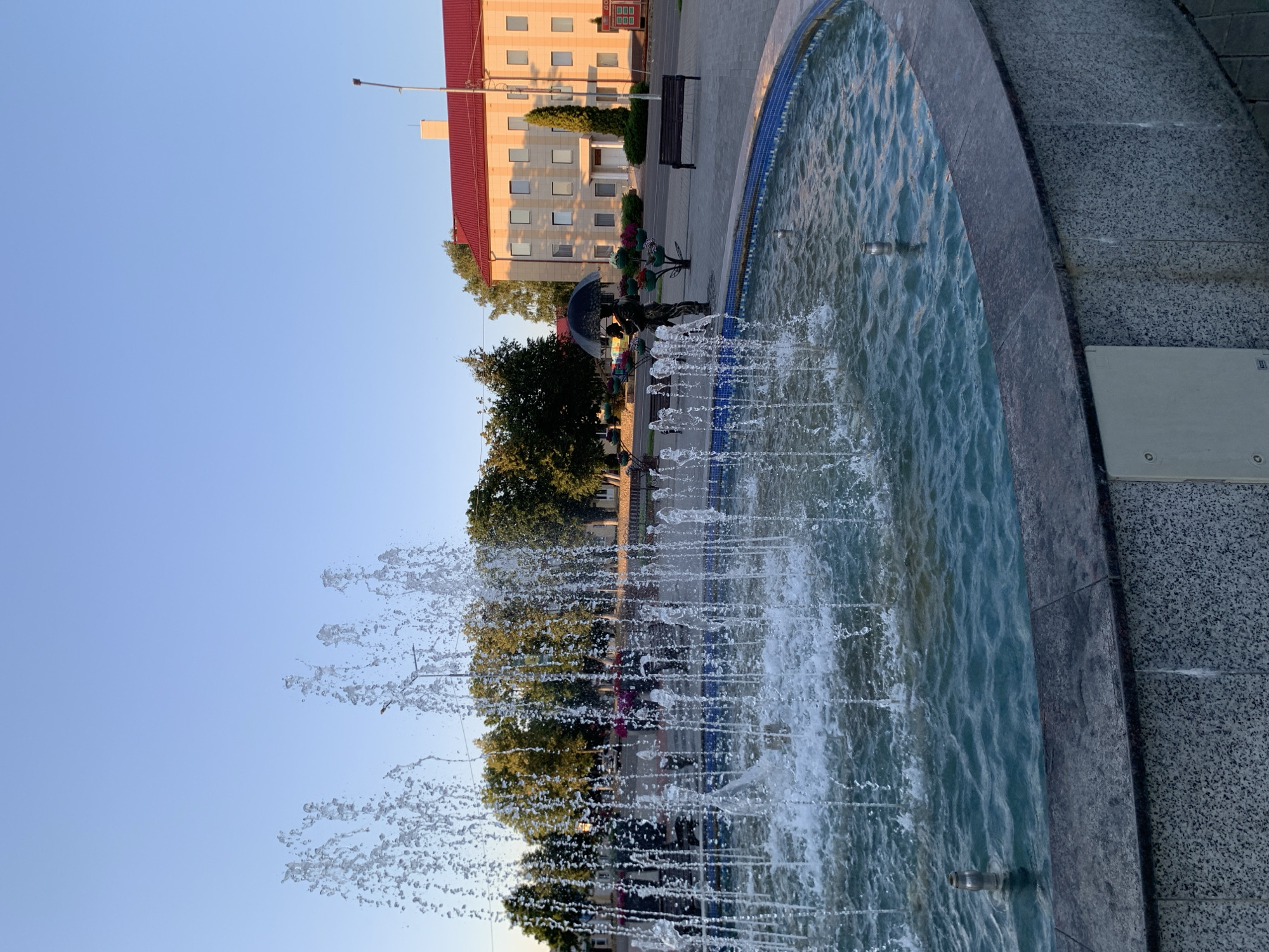 The image depicts a public square in Kapyĺ, Belarus, centered around a fountain. The fountain's water jets are prominently featured in the foreground, creating a sense of movement and freshness. In the midground, a bronze sculpture of a couple holding an umbrella stands near a decorative tree adorned with colorful flowers. Behind the sculpture, a light-colored building with a red roof dominates the background, suggesting it might be a public or administrative building. Trees line the perimeter of the square, and park benches are visible, indicating a space for public gathering and relaxation. The sky is clear and blue, with the warm light of what appears to be early morning or late afternoon sun illuminating the scene.