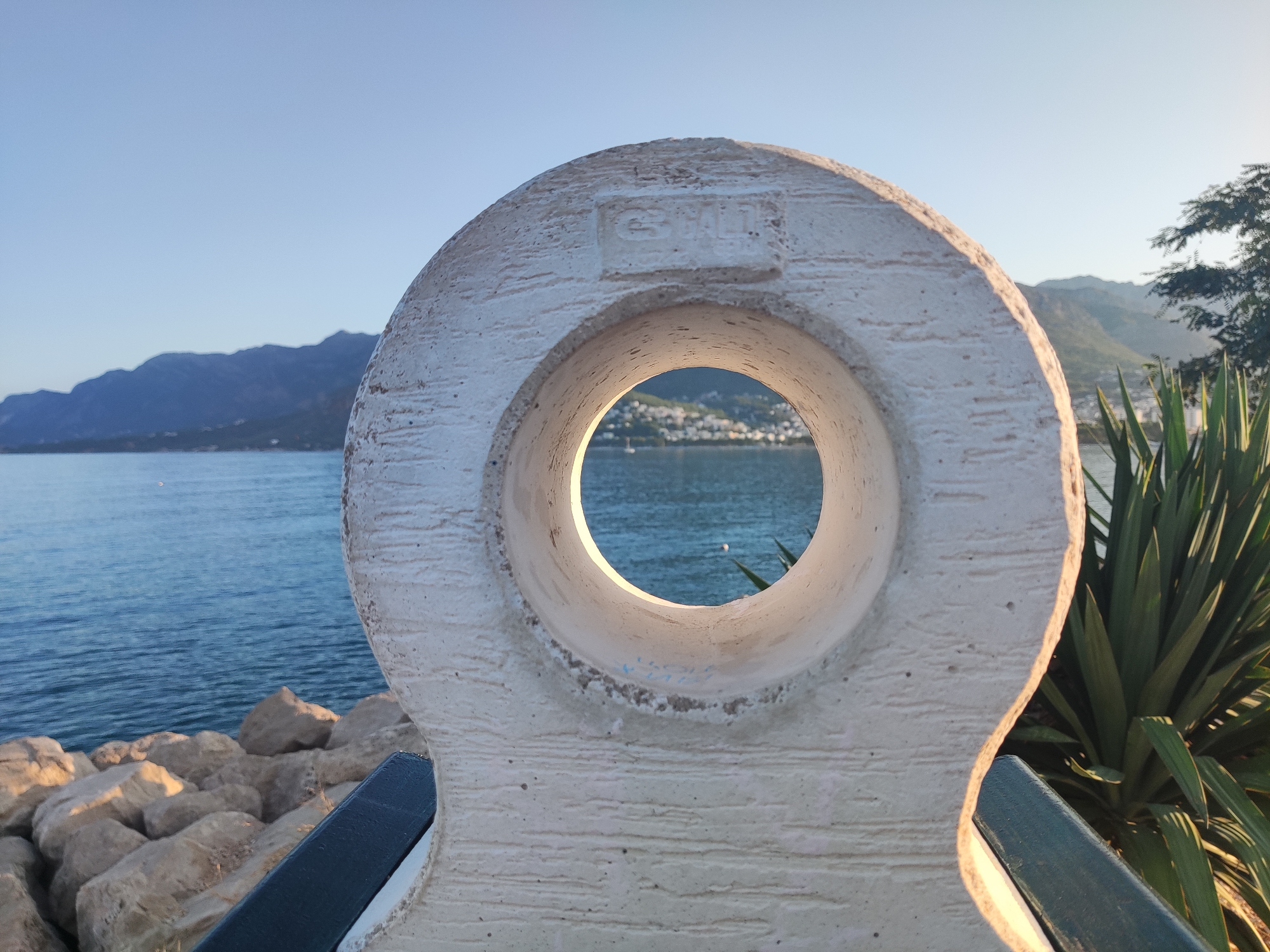 The image depicts a close-up view of a circular, concrete object with a hole in the center, placed in front of a scenic coastal landscape. The object, which appears to be a decorative element or a piece of art, is textured and light-colored. Through the hole, a body of water, likely the Adriatic Sea, is visible, with a distant town nestled along a hillside and mountains rising in the background. The sky is clear and blue, suggesting it is daytime.
To the left of the concrete object, a rocky shoreline is visible, with large, light-colored stones. To the right, lush green foliage, possibly a palm tree or similar plant, adds a natural element to the scene. The overall impression is one of a picturesque location, likely a public space or promenade, with a focus on the interplay between man-made structures and the natural environment.
The object in the foreground displays some embossed text within a rectangular area. Although partially obscured and weathered, the text appears to read "G. ŠAL." followed by other characters that are difficult to decipher. This inscription may indicate the artist or manufacturer of the object.
Considering the architectural style of the town visible in the background, characterized by buildings with red-tiled roofs and dense construction on a hillside overlooking the sea, and the presence of palm-like vegetation, this scene is strongly suggestive of a Mediterranean coastal town. The specific geographical context of Bar, Montenegro, known for its picturesque coastline, historical sites, and mountainous backdrop, aligns well with the visual elements presented in the image. The style of the concrete object and the overall ambiance are consistent with public art installations or maritime-themed elements often found in such coastal cities. Therefore, it is highly probable that this image was captured in the city of Bar, Montenegro, likely along its waterfront promenade or a similar public area.
