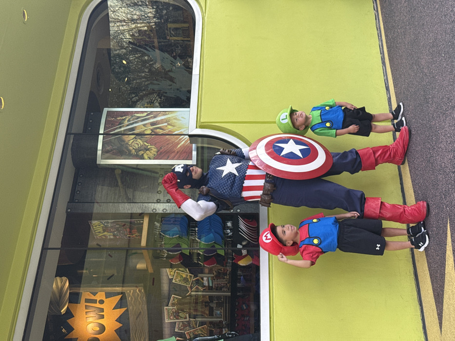 The scene captures three individuals posing outdoors in front of a lime-green building located in Marvel Super Hero Island at Universal's Islands of Adventure theme park in Orlando, Florida, United States of America.
A person dressed as Captain America stands centrally, facing slightly left. The individual wears the complete Captain America costume, including a blue mask with an 'A' emblem, a blue and red torso with a white star and red stripes, white sleeves with red gloves, blue pants, and red boots. A brown belt with pouches is visible. The right arm is bent with a clenched fist raised near the head, while the left arm holds a circular red, white, and blue shield with a white star in the center.
To the left of Captain America is a young male child dressed as Mario. He wears a red cap with a white 'M' emblem, a red shirt, blue overalls, black shorts, and black athletic shoes. His right hand is raised, touching the brim of his cap.
To the right of Captain America is another young male child dressed as Luigi. He wears a green cap with a white 'L' emblem, a green shirt, blue overalls, black shorts, and black athletic shoes. His arms are at his sides, and he is looking slightly upwards and to the right.
The background consists of a bright lime-green building facade with large, dark-framed windows. The left window displays a yellow "POW!" sign, an "X-MEN" comic book, and various retail merchandise. The right window features comic artwork depicting a powerful, muscular character in action, along with reflections of outdoor trees and distant structures. The ground beneath the individuals is dark pavement marked by a thin yellow line along the base of the building. The scene is illuminated by natural daylight.