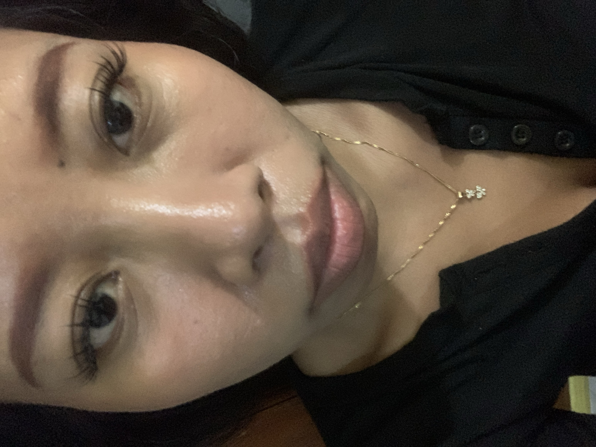 The image is a close-up, overhead selfie of a woman with dark hair and light brown skin. She is wearing a black, button-down shirt and a delicate gold chain necklace with a small, bejeweled pendant. Her makeup includes defined eyebrows, false eyelashes, and lip gloss. The lighting in the image is bright, causing some glare on her forehead and cheeks. The background is dark and out of focus, suggesting an indoor setting. The specific location within Jajag, Indonesia cannot be determined from the image.