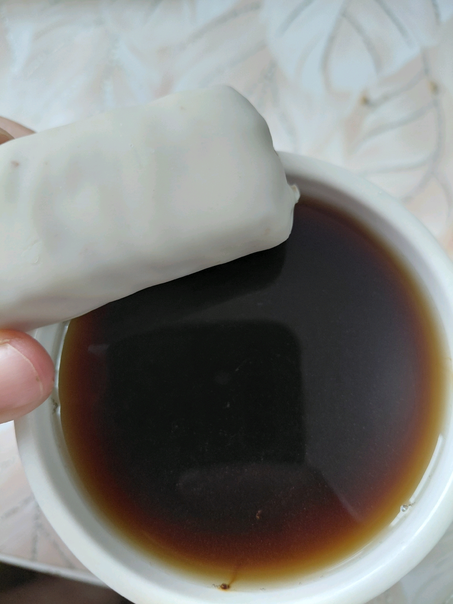 A close-up shot shows a person's thumb and index finger holding a white rectangular object, possibly a food item, partially dipped into a white cup filled with a dark liquid, likely coffee or tea. The background consists of a patterned surface, possibly a tablecloth or placemat, with a light, intricate design. The scene appears to be indoors, with natural lighting. The image does not contain any elements that would suggest it is located in Malinovka, Russia.