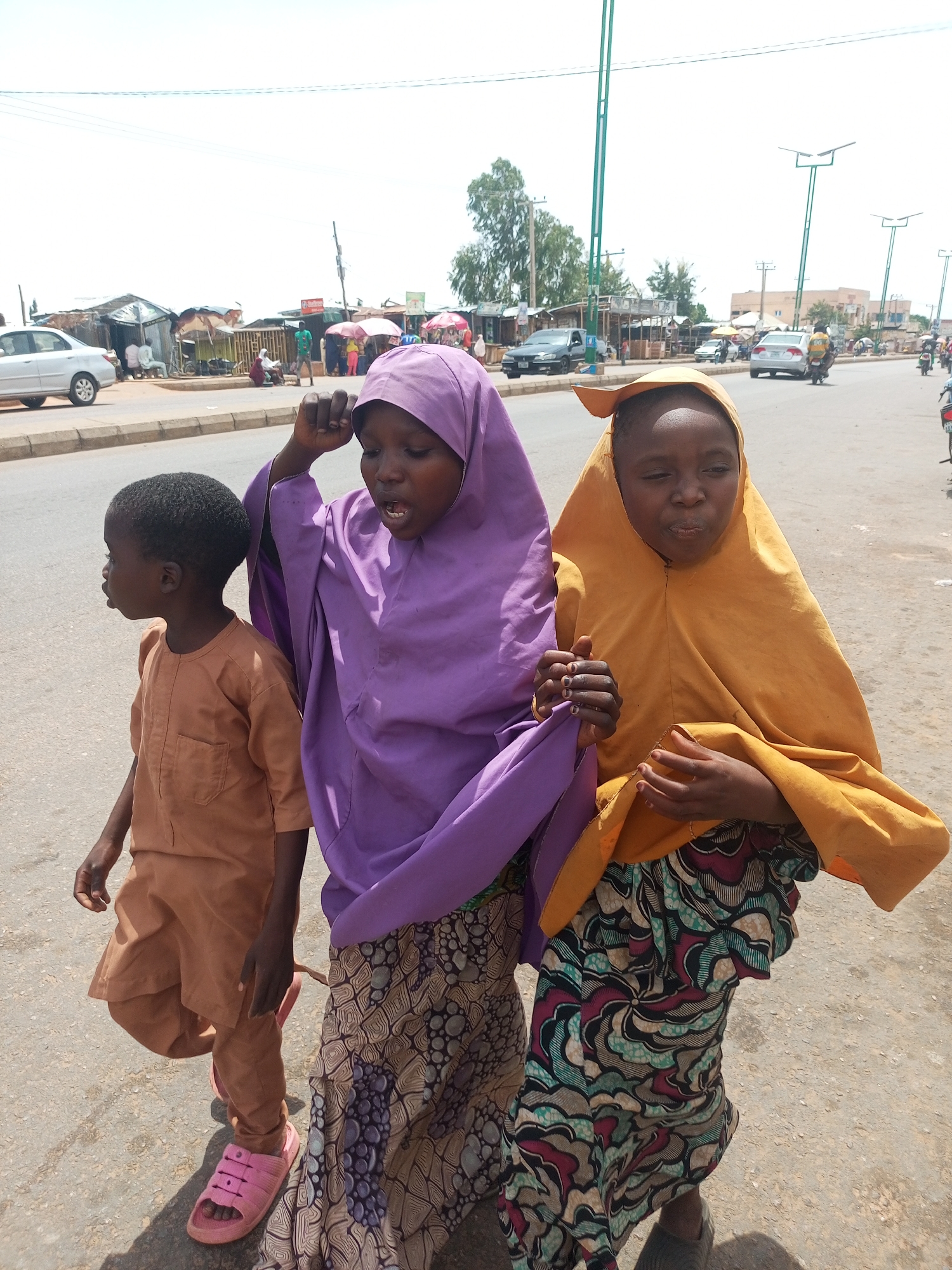 In Gombe, Nigeria, three children walk along a paved road, likely a street in the city, with shops and stalls lining the left side of the road. The child on the left, a boy wearing a light brown tunic, walks ahead. To his right, two girls, both wearing headscarves, walk together. The girl in the middle wears a purple headscarf and a patterned skirt, while the girl on the right wears a yellow headscarf and a colorful, patterned skirt. The girls appear to be engaged in conversation, with the middle girl gesturing with her raised hand. The background shows a typical urban scene, with several vehicles, including cars, a motorcycle, and a truck, driving on the road. Overhead, power lines and streetlights suggest an active commercial or residential area. The ground is a paved road with some loose gravel and debris. The sky is bright and overcast, indicating a daytime scene.