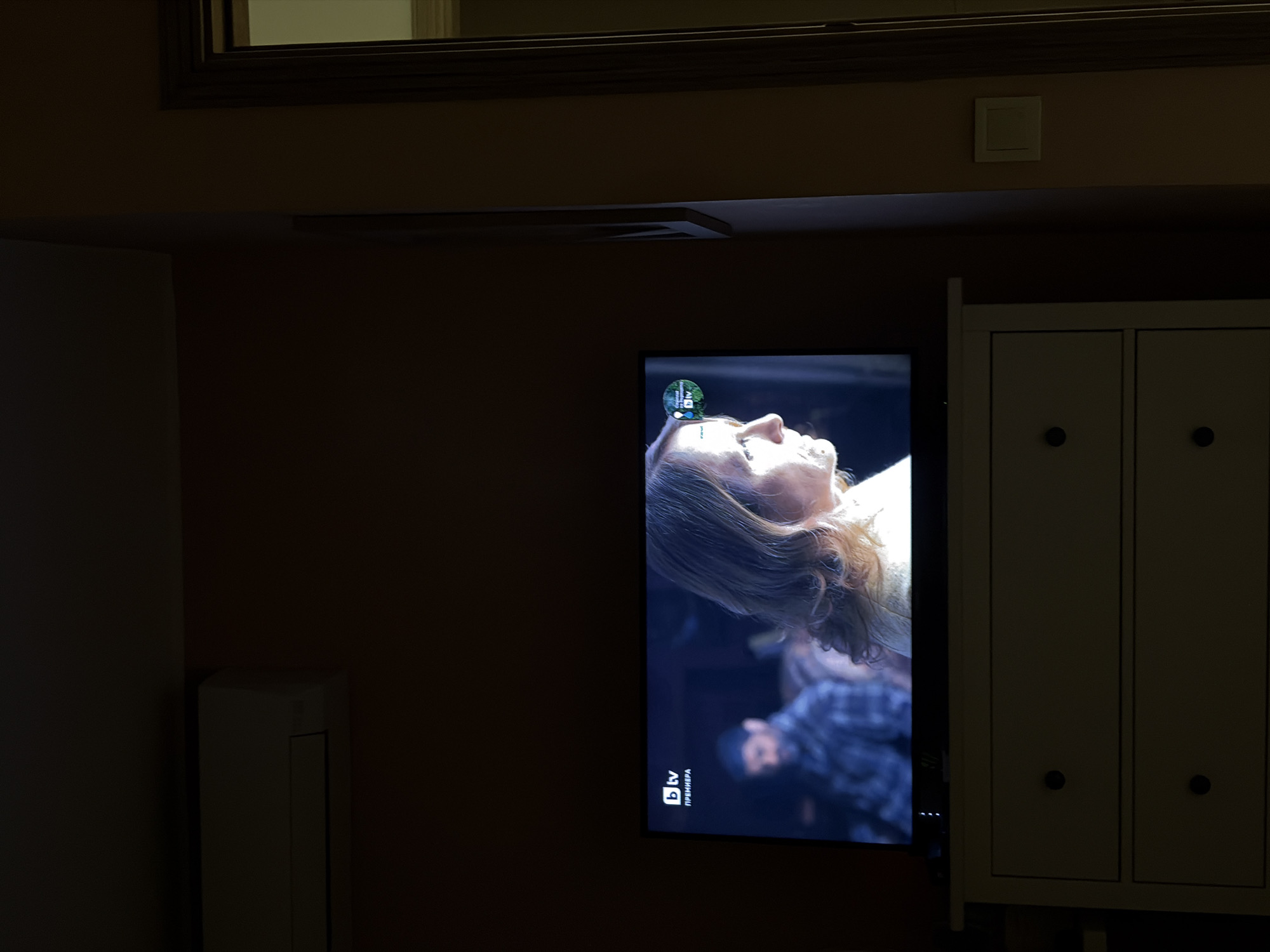 The image shows a darkened room with a television mounted on the wall, displaying a woman's face. To the right of the television is a light-colored cabinet with two doors. A white air conditioning unit is visible to the left of the television. A dark wooden frame is visible near the top of the image. The television is tuned to bTV, a Bulgarian television channel. No information suggests the location is in Burgas, Bulgaria.