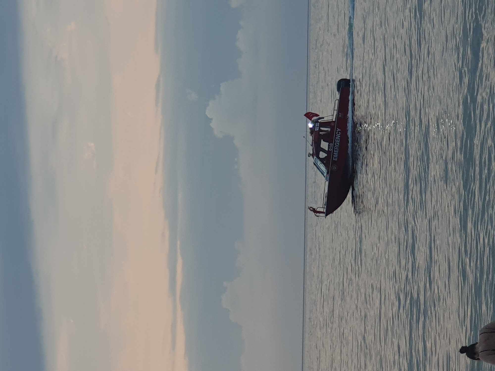 An emergency response boat, marked "EMERGENCY" in white lettering, is cruising on the calm sea near Batumi, Georgia. The boat is red and has a light on the roof and a flag at the stern. Two people are visible on the boat, and another person is partially visible in the foreground in the water. The sky is cloudy with hints of pink and gray. The sea is flat and reflects the sky and the boat.