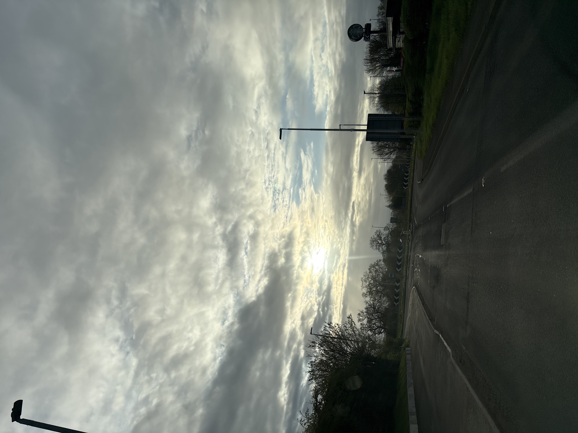 The image depicts a road in Oswestry, United Kingdom, on a cloudy day. The road stretches into the distance with markings and street furniture on either side. Trees and vegetation line the edge of the road. The sky is predominantly covered with clouds, with the sun peeking through in the distance.