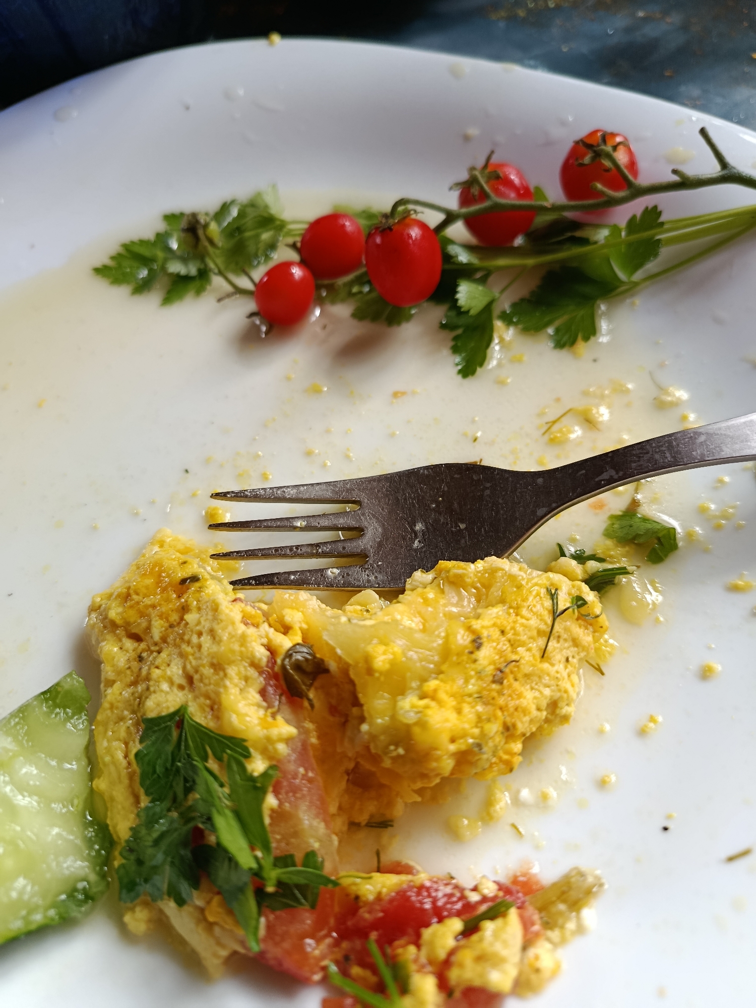 The image shows a white plate with the remains of a meal, likely an omelet, with some scattered ingredients. A fork is placed on the plate, partially covering the food. There are several cherry tomatoes on a stem with parsley, and a slice of cucumber is visible on the left side of the plate. The plate itself is on a dark, textured surface, possibly a table. The scene suggests a recently finished meal. There is no indication of the city of Верея, Russia in the image.