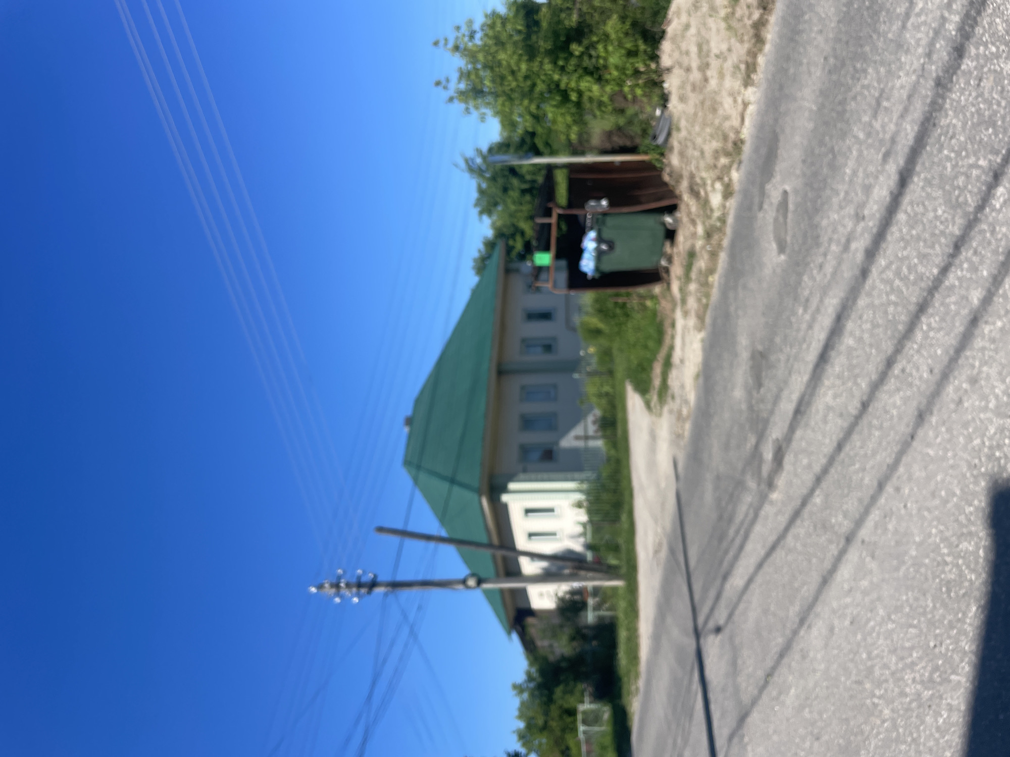 The image depicts a rural scene, likely in the village of Krasnye Baki, Russia. A modest, light-colored house with a prominent green, gabled roof stands on the right side of the frame. In front of the house and slightly to the right, a green dumpster is situated within a rudimentary metal enclosure. The dumpster appears to contain some white plastic bags. A dirt path leads from a paved road towards the house. A power pole with electrical lines running overhead is positioned to the left of the house. The surrounding area is characterized by lush green trees and vegetation, with a clear, bright blue sky overhead. The road itself is asphalt and shows signs of wear, including some shallow depressions. The overall impression is one of a quiet, sun-drenched day in a residential area outside of a major urban center.