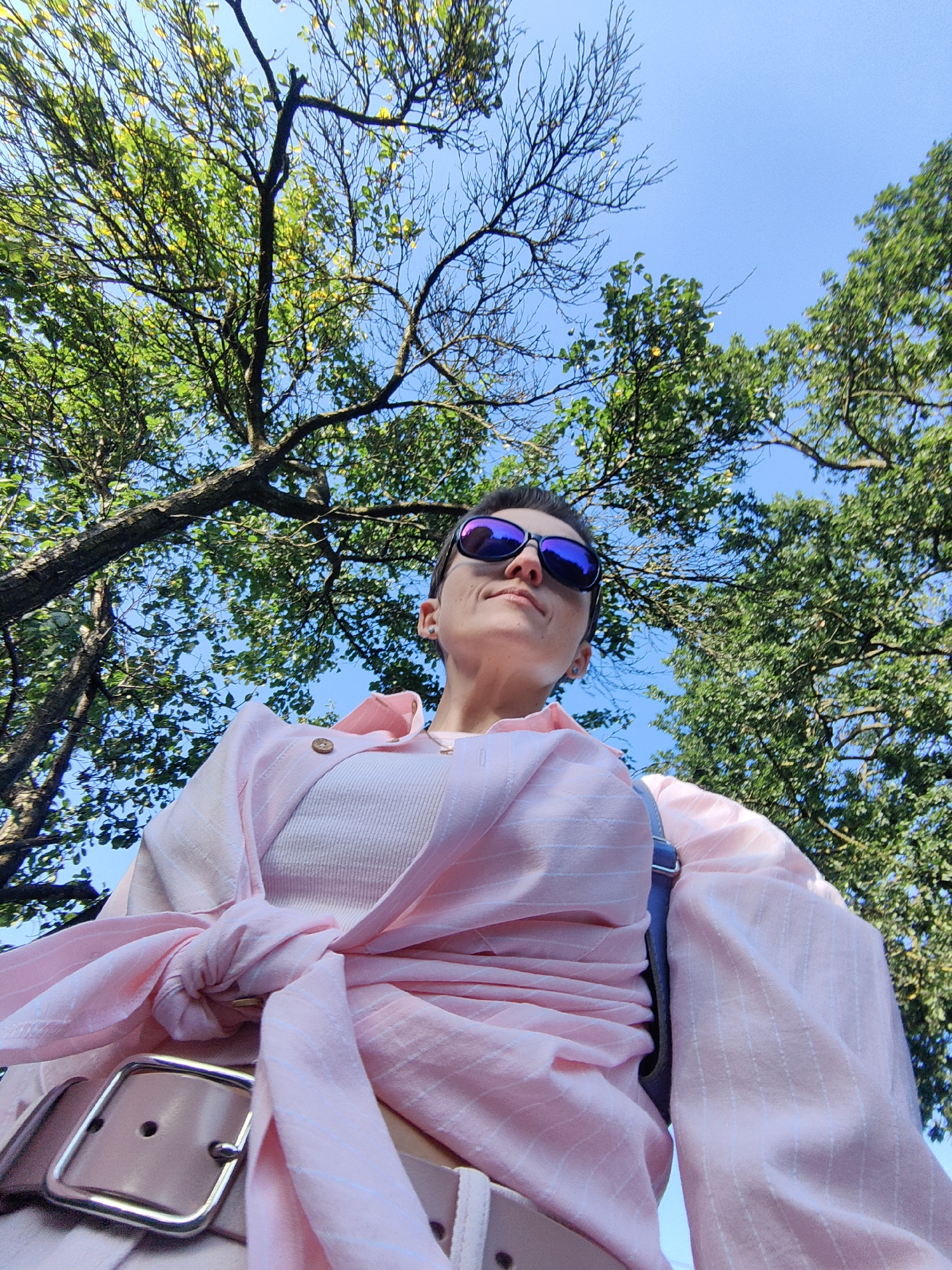 The image, taken from a low angle, shows a person wearing sunglasses and a pink outfit, looking upwards. The person is outdoors, surrounded by lush green trees and a clear blue sky. The scene appears to be set in a park or natural area within the city of Fanipal, Belarus. The person's attire suggests a casual or fashionable outing. The sunlight illuminates the foliage and casts subtle shadows, indicating it is daytime.