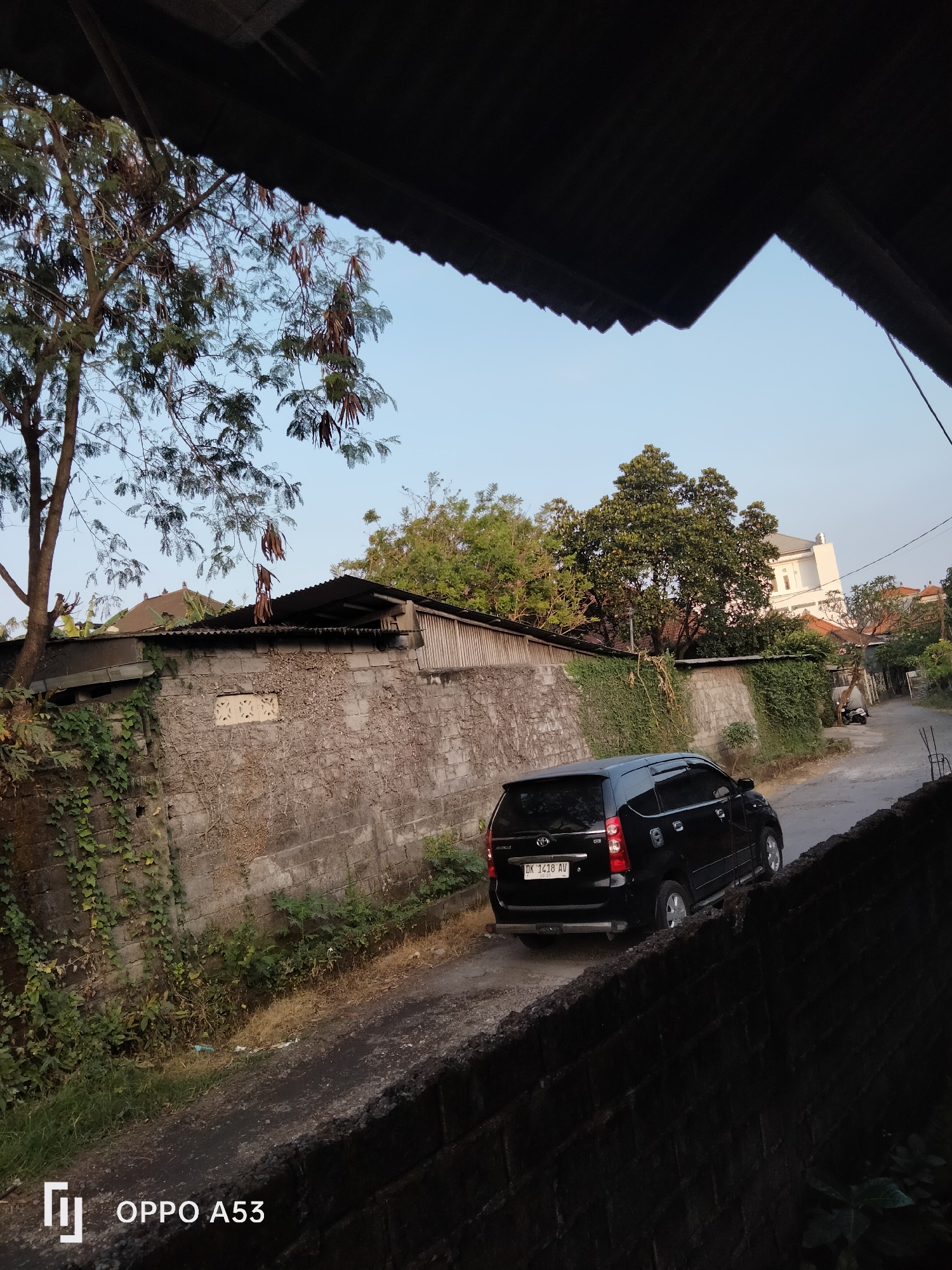 The image depicts a residential street scene in Denpasar, Indonesia, likely in the late afternoon or early morning given the soft, warm lighting. A black minivan, possibly a Toyota Avanza, is parked on the right side of a narrow, unpaved road. The vehicle has a license plate that reads "DK 1418 AV". To the left of the road, a weathered stone wall, partially covered in ivy and dry vegetation, runs along the property line. Behind the wall, the roofs of several houses are visible, with one white building standing out in the background. Trees with sparse foliage and some dry seed pods hang over the scene from the left. The foreground is dominated by a dark, textured surface, possibly another wall or a shaded structure, with the "OPPO A53" watermark at the bottom left corner of the image. The sky is a clear, pale blue. There are no people visible in the image, and no specific activity is taking place other than the parked vehicle. The overall impression is one of a quiet, everyday urban or semi-urban environment in Denpasar.