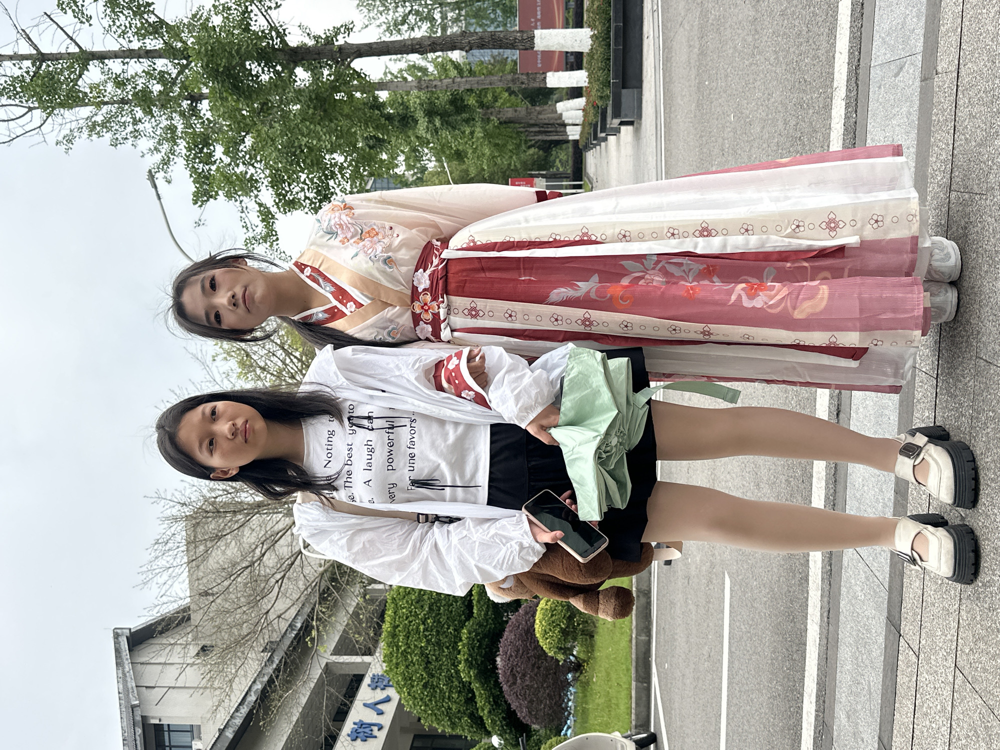 Two young women are posing outdoors. The woman in the background is wearing a traditional Chinese dress with floral patterns in shades of red, white, and peach. The woman in the foreground is dressed in a white graphic tee with black shorts and a white jacket. She is holding a cell phone in one hand and a folded green umbrella in the other, with a small teddy bear hanging from her bag. They are standing on a paved walkway with trees and greenery in the background. In the distance, there is a building with Chinese characters visible. The location is Jiangjin, China.