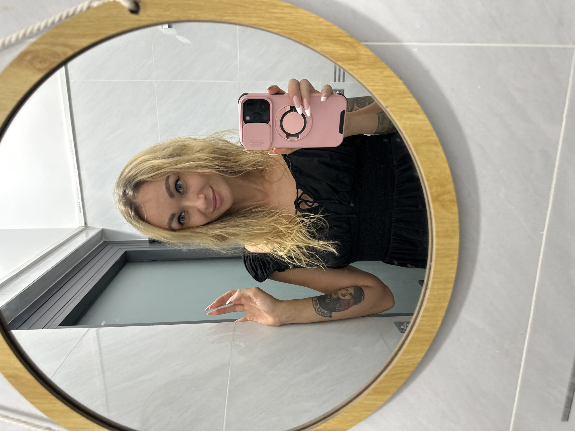 A mirror selfie captures a young woman with long, wavy blonde hair and striking blue eyes, looking directly at the camera with a gentle smile. The image is rotated 90 degrees counter-clockwise. She is holding a smartphone in a pink case, featuring a black camera module and a circular pink pop socket/ring holder. Her nails are long and manicured with white French tips. She wears a black top or dress with smocked detailing and short puffed sleeves. A prominent tattoo, possibly depicting two faces, is visible on her left forearm, which she rests on a light-colored surface within the reflection.
The selfie is taken in a round mirror with a light wooden frame and a visible rope hanger at the top. The reflection reveals a room with light grey tiled walls. A dark-framed window or partition is also visible in the background reflection, suggesting a space beyond. This scene is likely set in Mũi Né, Vietnam. The overall impression is one of casual confidence.