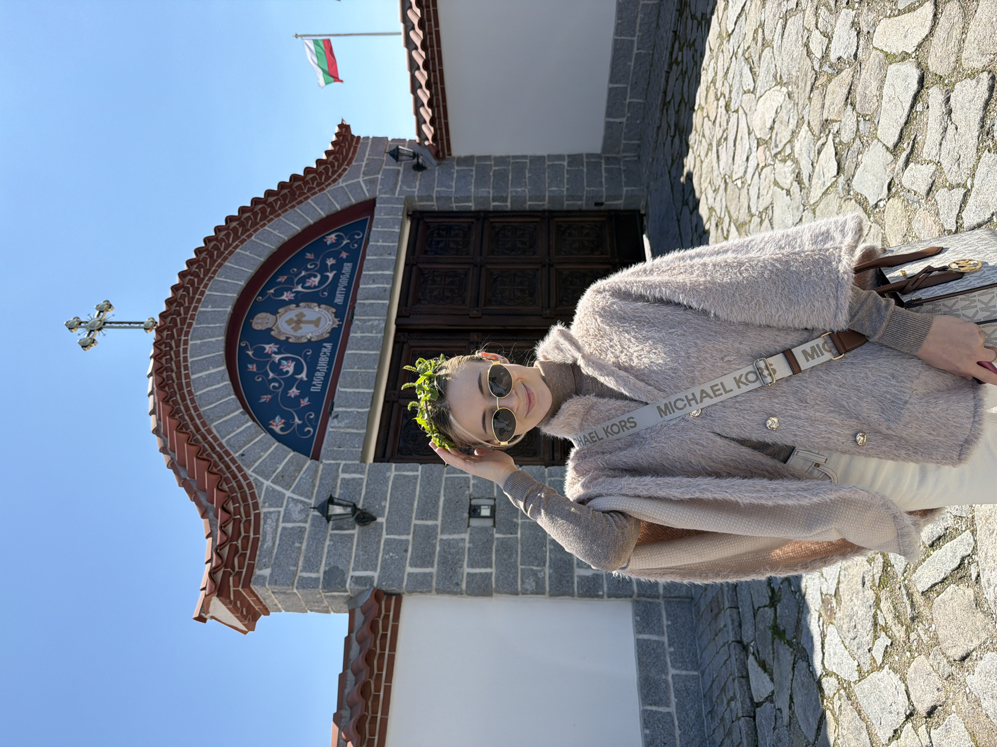 A woman wearing a flower crown and sunglasses stands in front of a stone building with a tiled roof and a Bulgarian flag waving above. The building has a wooden door and an inscription above it. The woman is wearing a brown fuzzy coat, a gray turtleneck, and a white Michael Kors purse. The ground is paved with stones. The photograph was taken during the day in the city of Plovdiv, Bulgaria.
