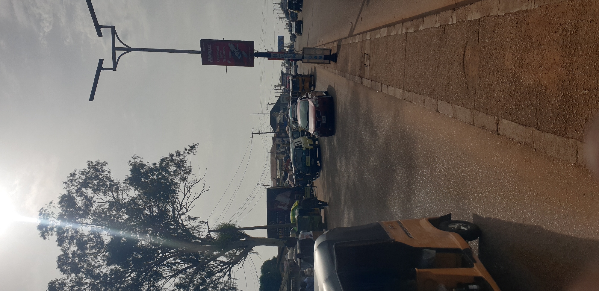 An outdoor, rotated photograph captures a busy street scene in Sabon Tasha, Nigeria, under a bright, sunny sky during the daytime. The perspective looks down the road, with a paved surface for vehicles and a raised, unpaved sidewalk made of gravel or dirt on the right.
In the foreground, a yellow and black tricycle (likely a Keke Napep or auto-rickshaw) is visible on the right side of the road. Further down the street, a line of vehicles includes a prominent red car, as well as several yellow and green vehicles, suggesting commercial use like taxis or public transport. People are seen near these vehicles, though their features are indistinct.
On the left side of the road, a tall utility pole with a distinctive multi-armed crossbar structure stands, supporting power lines that crisscross the frame. Attached to this pole is a large billboard, oriented perpendicularly to the road. Visible text on the billboard includes "Globacom" (a telecommunications company logo) and "Unlimited Free Talktime," advertising a service. Beneath the billboard, a person in a bright green top is partially visible.
A large, leafy tree dominates the far left of the image, with bright sun glare emanating from behind its branches, indicating the sun's position. More buildings and vehicles line the street in the distance, creating a sense of an active urban or semi-urban environment. The sky is bright but has some atmospheric haze.