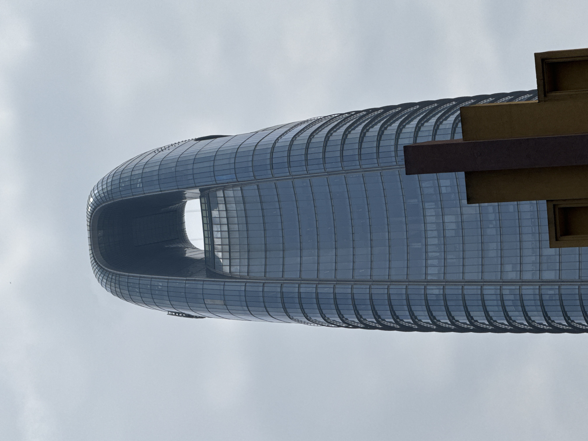 The image displays a prominent view of the Guangzhou Circle building, a distinctive modern architectural landmark located in Haizhu District, China. The photograph captures the building against a soft, overcast sky, highlighting its unique design.
The skyscraper presents a circular or elliptical form with a large, hollow void in its center, resembling a jade disc or an ancient coin. Its exterior is predominantly clad in reflective glass panels, giving it a sleek, blue-gray appearance that subtly mirrors the diffused light of the cloudy day. The facade is characterized by horizontal lines and segmented patterns, indicative of its complex structure.
On the right side of the frame, angular, earth-toned architectural elements are partially visible in the foreground. These brown and dark red structures, possibly part of an adjacent building or a decorative feature, offer a contrasting texture and color against the smooth glass of the main tower.
The sky is a uniform light gray, suggesting an overcast or cloudy day, which provides a neutral backdrop for the building. There are no people visible in the image, nor any discernible text. The photograph emphasizes the building's monumental scale and its bold, futuristic architectural statement within an urban setting.