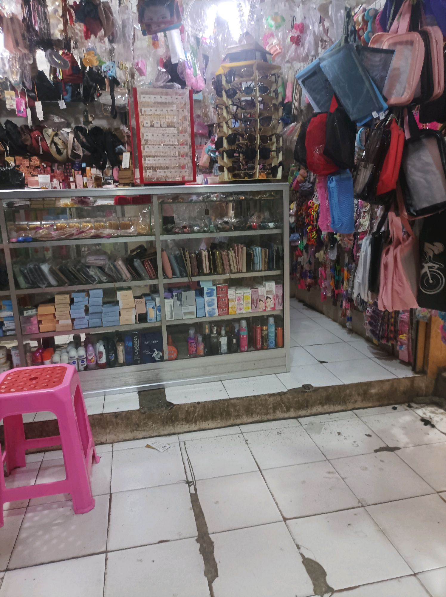 The image depicts a market stall in Sumuragung, Indonesia, filled with various goods. In the foreground, a bright pink plastic stool sits on a tiled floor. Behind the stool, a glass-fronted display cabinet showcases a variety of items, including what appear to be toiletries, makeup, and accessories. The shelves are neatly organized with boxed products.
To the right of the display cabinet, more merchandise is visible, including handbags, pouches, and what looks like costume jewelry. The stall is densely packed with items, suggesting a busy marketplace environment. In the background, the ceiling and upper walls are visible, also adorned with hanging goods, indicating the extensive nature of the merchandise. The overall impression is of a well-stocked stall in a bustling local market.