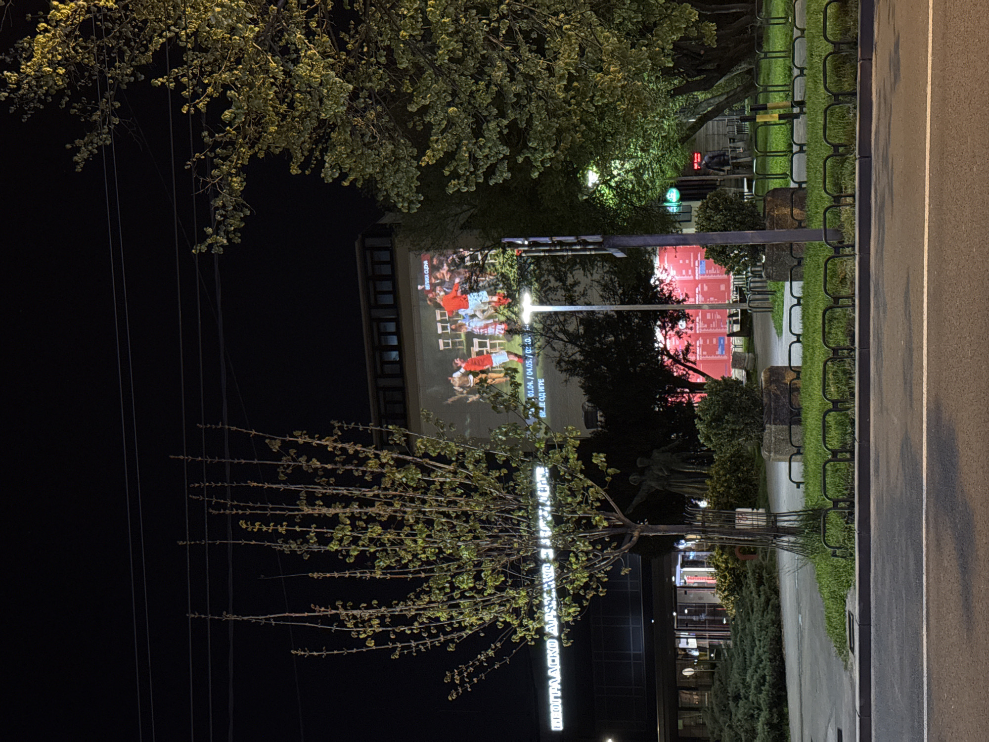Nighttime shot in Belgrade, Serbia, depicting a cityscape with prominent buildings, trees, and a street. A building on the left is illuminated with a large projected image of a group of people. To the right, another building displays pinkish-red lights. The street has a bicycle rack and green grassy areas. An illuminated tree is in the foreground. Overhead power lines are visible in the dark sky. The scene suggests an urban environment at night, with the illuminated buildings serving as focal points.