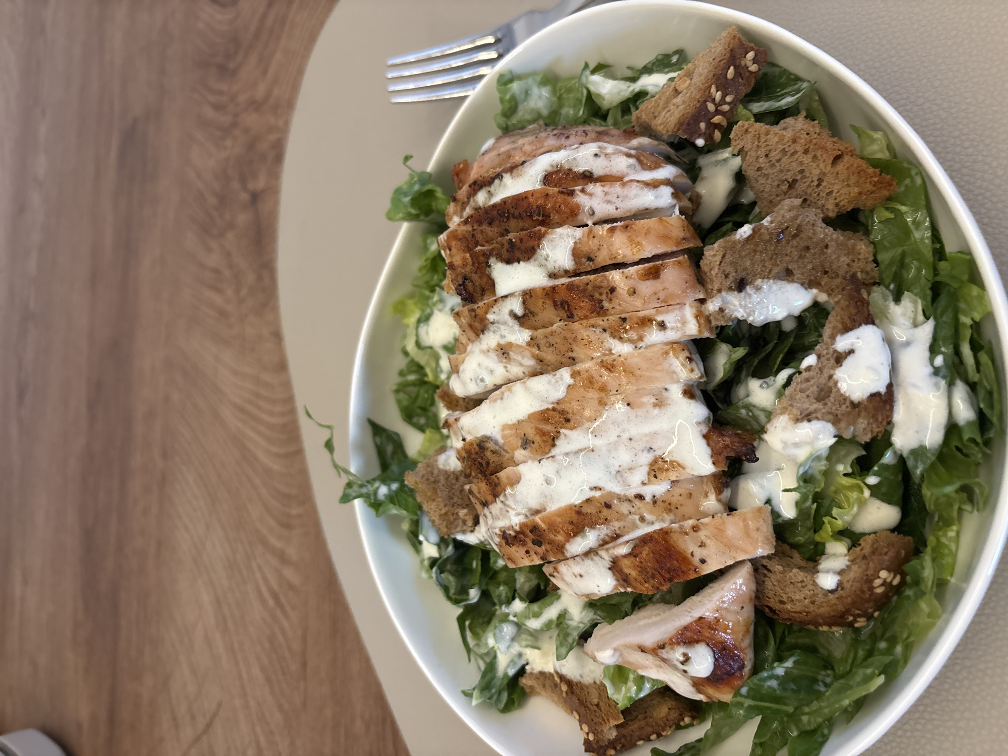 A close-up, eye-level shot captures a delicious-looking Caesar salad with grilled chicken and croutons in a white bowl. The salad is generously drizzled with creamy dressing, and the chicken is sliced and arranged attractively on top. To the right of the bowl, a silver fork rests on a light brown placemat, which is on a wooden table. The background is blurred, showing a hint of the wooden table's texture. The overall impression is one of a simple, yet satisfying meal. This image was taken in Viernheim, Germany.