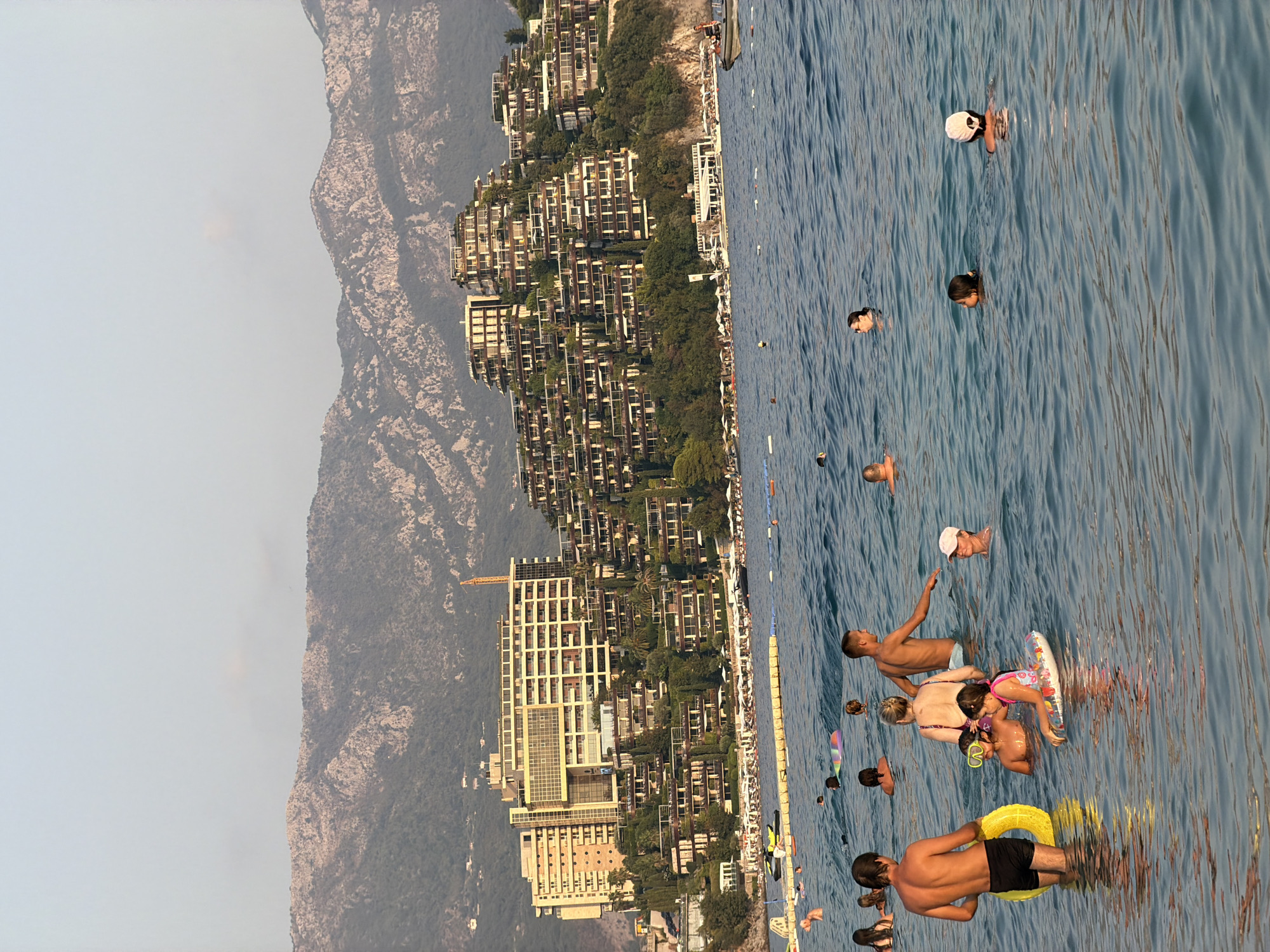 The image shows a crowded beach and the sea in front of a resort complex in Budva, Montenegro. In the foreground, several people are wading and swimming in the clear blue water. Some are using floatation devices, including a yellow ring and a patterned inflatable.
In the mid-ground, a long, yellow floating barrier separates swimmers from a section of the beach. Beyond the barrier, the beach is lined with numerous white umbrellas and lounge chairs. Several boats and a jet ski are visible near the shore.
The background features a multi-tiered resort complex built into the hillside. The buildings are modern, with many balconies and terraces, and are surrounded by lush greenery. A construction crane is visible behind one of the buildings, suggesting ongoing development. Towering mountains, with rugged, rocky slopes, rise behind the resort, creating a dramatic backdrop under a hazy sky. The overall scene depicts a popular tourist destination during daylight hours.