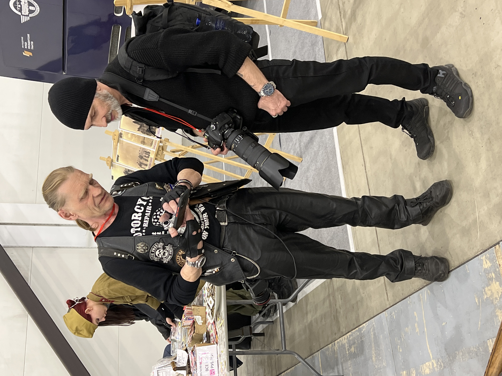 Here is a description of the image:
The image captures two men standing at an exhibition or event. The man in the foreground is dressed in black leather pants, a black t-shirt with motorcycle-themed graphics, and a leather vest adorned with patches. He also wears fingerless gloves and a watch. He's looking at a table laden with merchandise, possibly taking a photograph with a smartphone. To his left, a person wearing a khaki-colored hat and jacket is partially visible.
Behind him, a second man is dressed entirely in black, including a beanie. He has a DSLR camera with a large lens slung around his neck and is looking down in the same direction as the first man, potentially taking a photograph.
The setting appears to be an indoor event, with tables displaying various items for sale. In the background, there is a display featuring images on small easels and part of a vehicle. The floor is light-colored and appears to be a polished surface.
The image does not offer enough information to identify a specific city or to determine the location as "Unknown, Unknown."