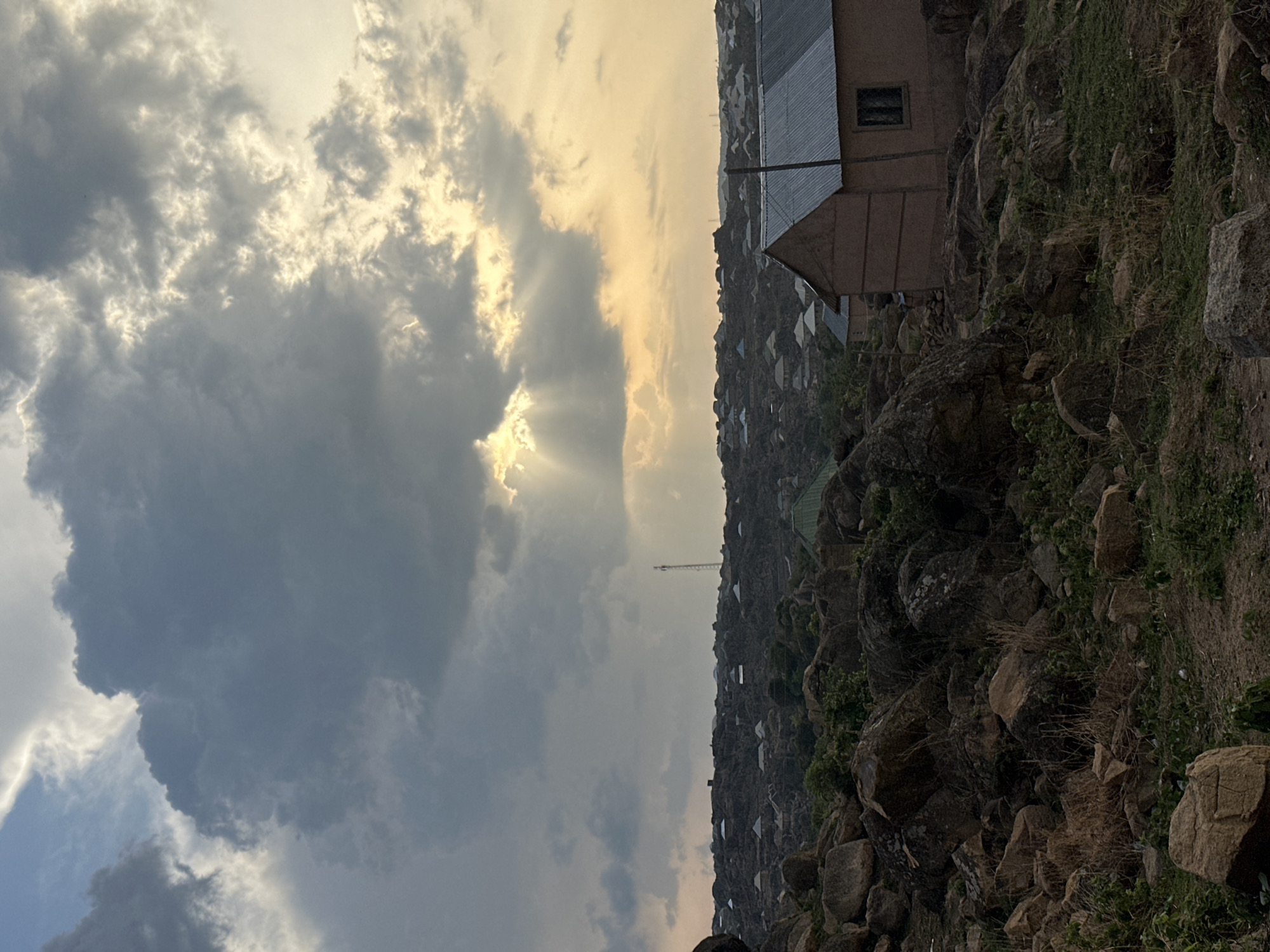 The image shows a landscape in Jos, Nigeria. The sky is filled with clouds, with sunlight breaking through in some areas. On the right side, there is a rocky terrain with some vegetation and scattered buildings. The buildings are simple, with metal roofs. A tall antenna is visible in the distance.