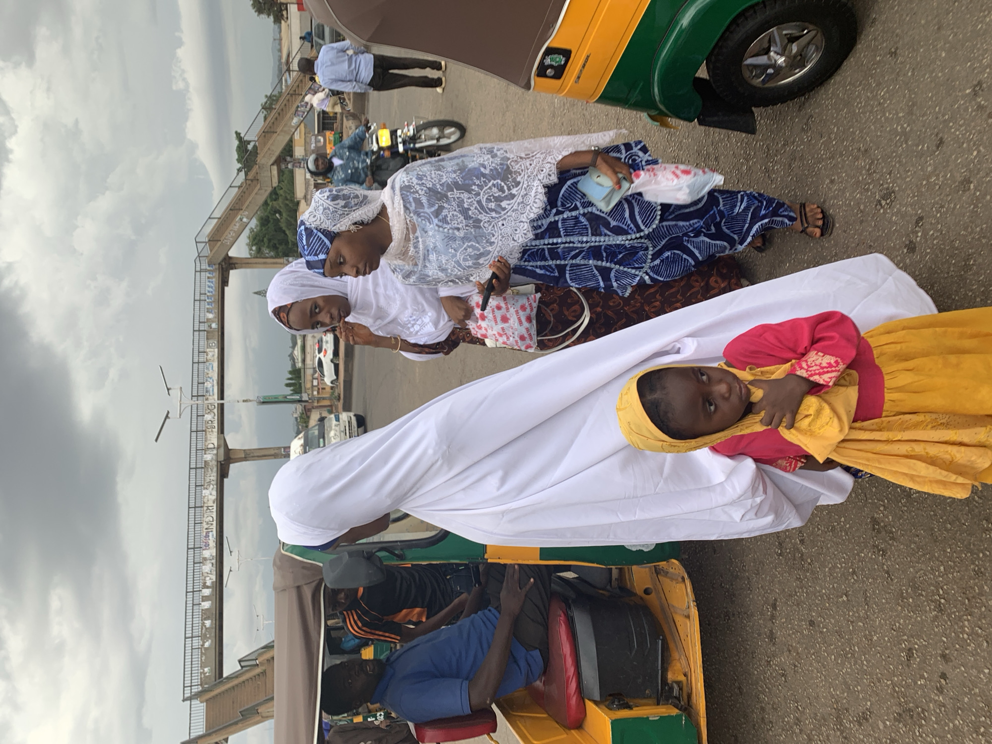 The image shows a scene on a street in Gura Top, Nigeria. Two women wearing headscarves and traditional patterned dresses are standing near a yellow and green auto rickshaw. One of the women carries a small purse and a plastic bag. A young child wearing a yellow and pink dress and a yellow headscarf is being held, swaddled in a white sheet, on top of the rickshaw. Inside the rickshaw, a driver wearing a blue shirt and another person wearing black can be seen. In the background, there is a pedestrian bridge, and other vehicles are passing by. The sky is overcast.