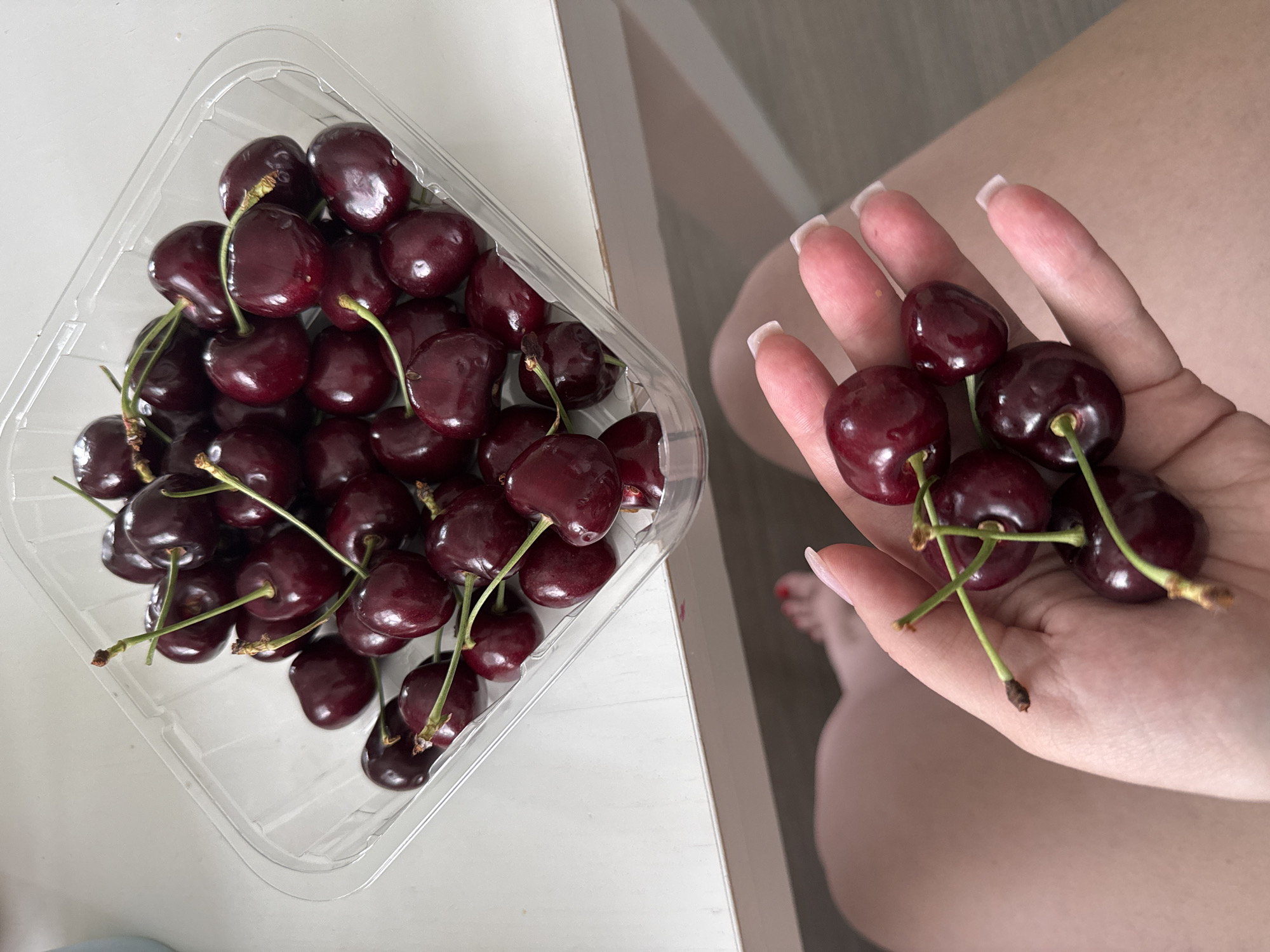 An overhead shot shows a person's hand holding a small cluster of ripe cherries, with stems attached, over their lap. The person's fingernails are manicured with a white polish. In the upper portion of the frame, a clear plastic container is filled with more cherries. The background consists of a light-colored surface and a hint of what appears to be legs. The scene is indoors, in what is presumed to be a domestic setting. The lighting is bright and natural, suggesting it was taken during the daytime.
