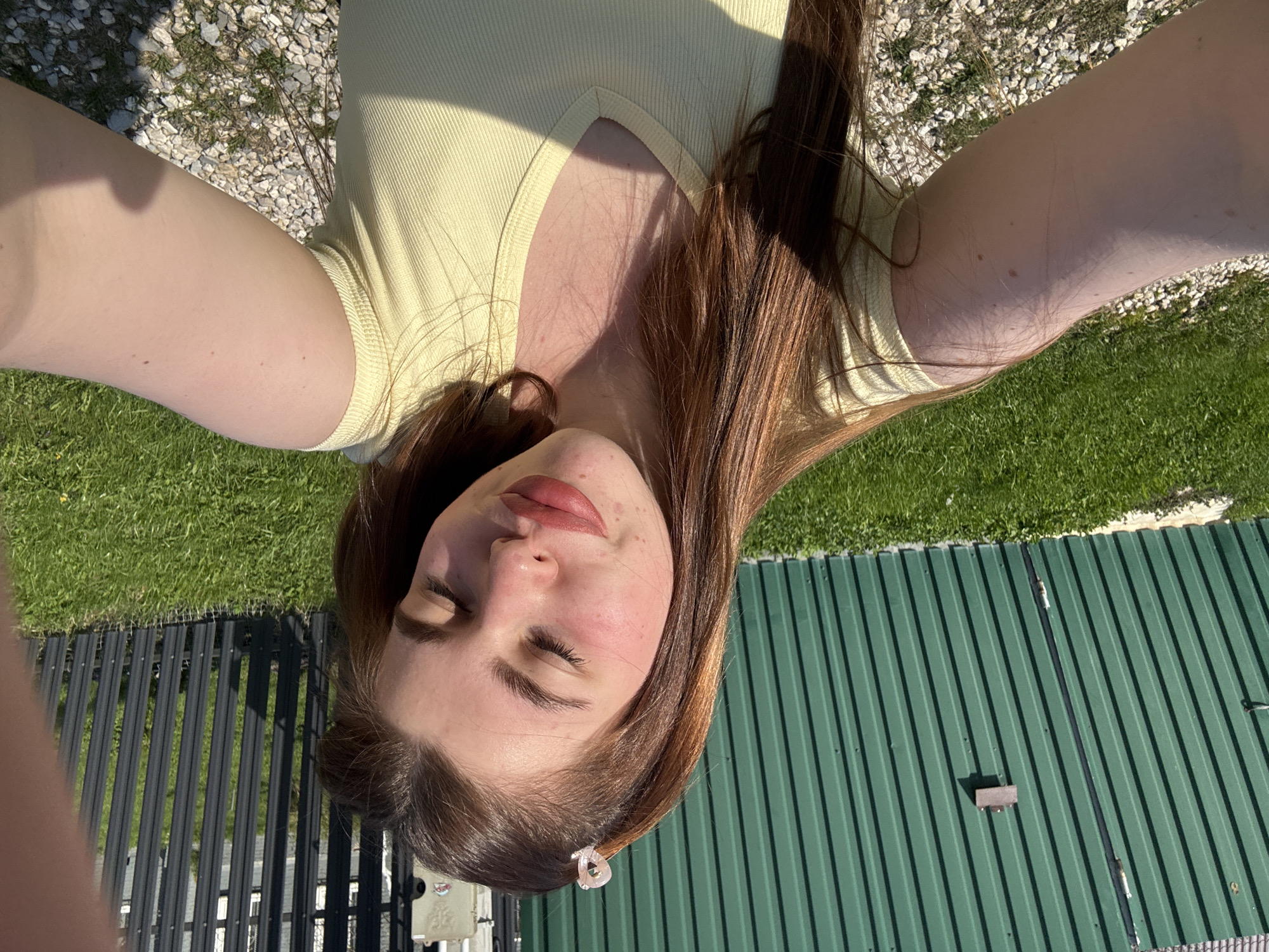 A selfie of a young woman with closed eyes, long auburn hair, and a pale yellow ribbed top is centered in the frame. The setting appears to be outdoors, with a combination of gravel, grass, and green corrugated metal in the background. A black metal fence is also visible in the lower-left portion of the image. The image is rotated counter-clockwise. The location cannot be verified as Шевлягино, Russia, based on the image alone.