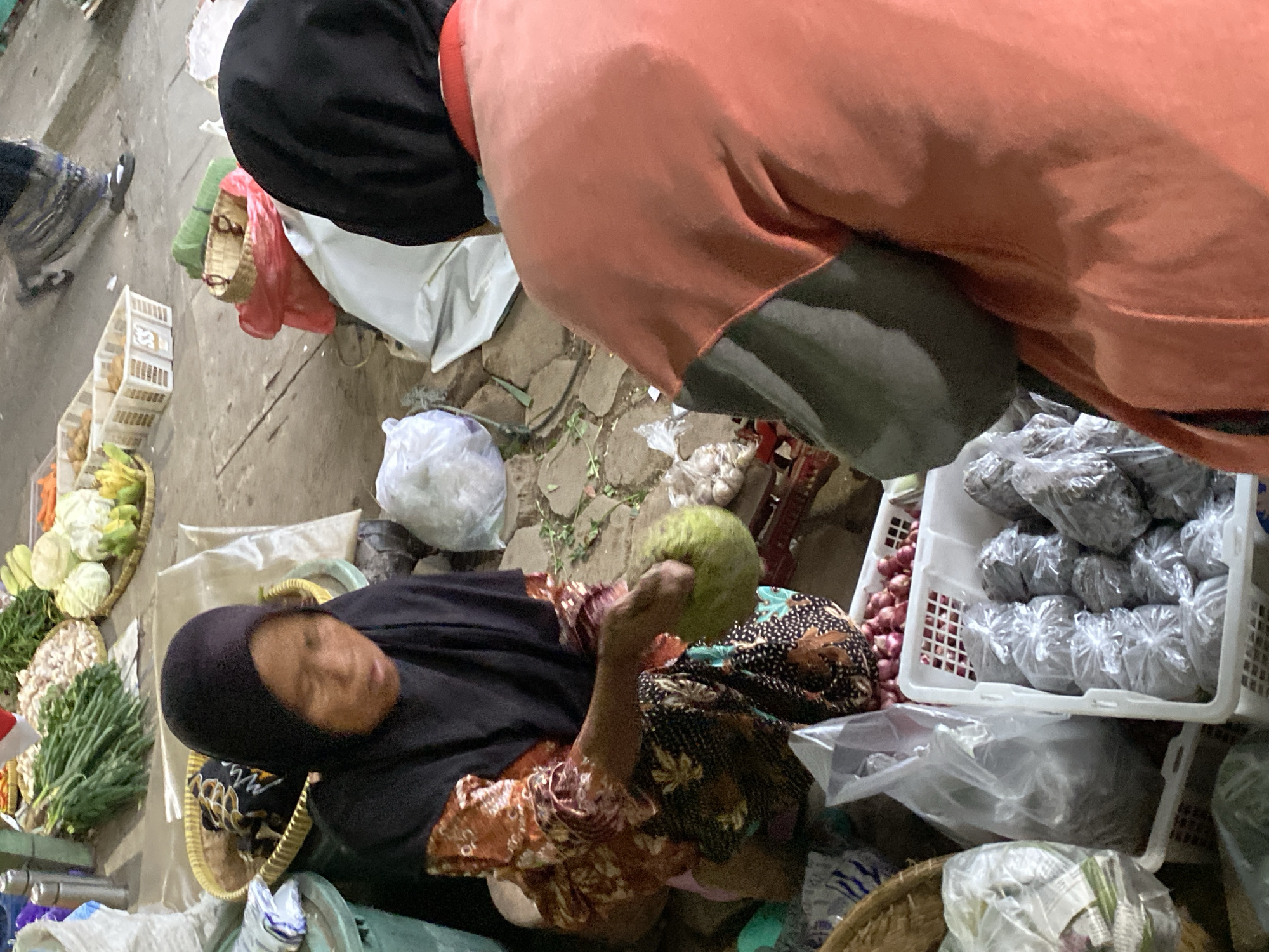 At a street market stall in Ambarketawang, Indonesia, two individuals are visible. In the foreground, a person wearing an orange and grey sweatshirt faces away from the camera. To their left, a person wearing a black hijab and a patterned orange and black top is seated, holding a large green fruit. They are interacting with each other. Various market goods, including produce in baskets and bags, are arranged around them. The ground is paved with rough stones.
