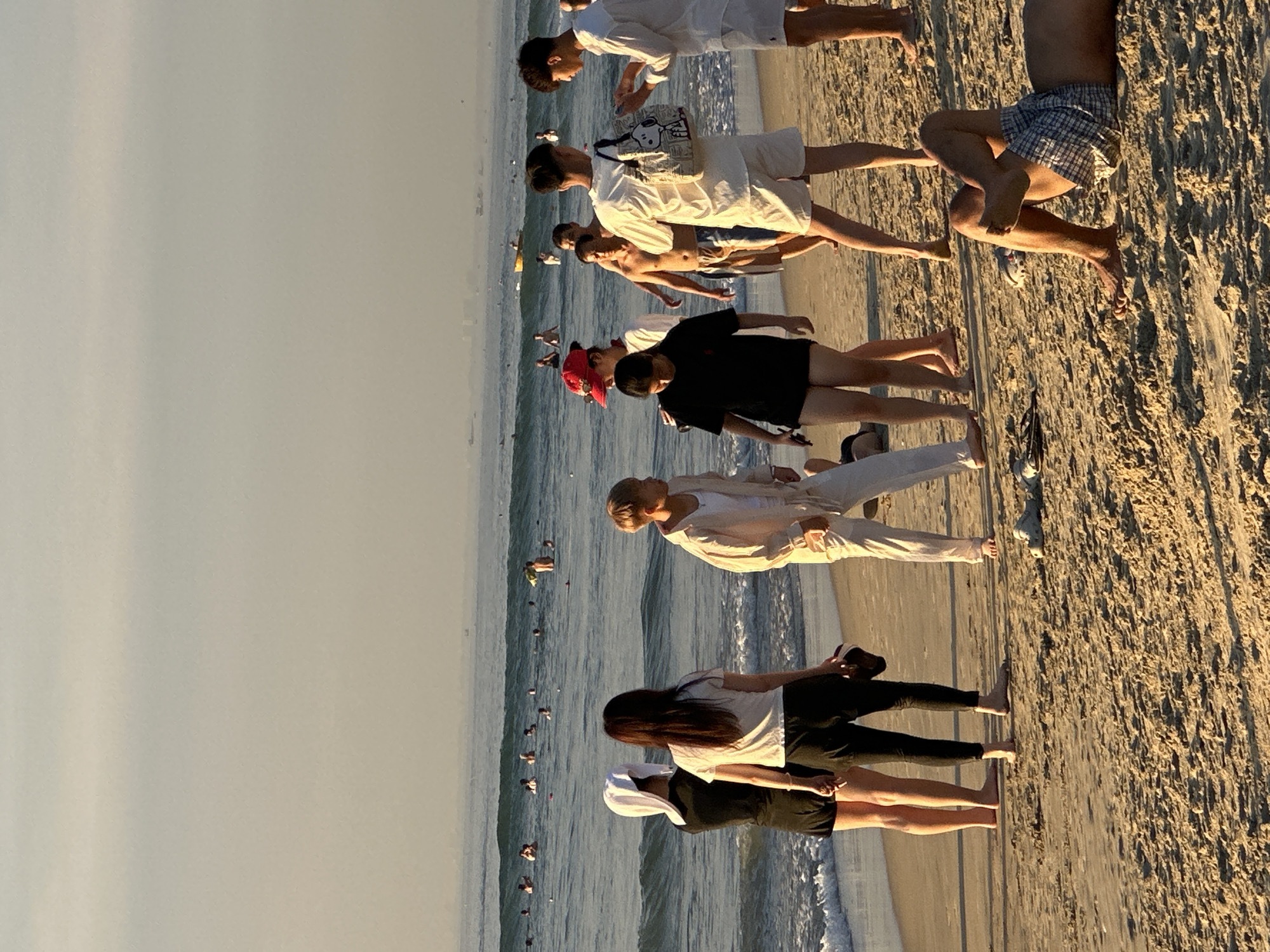 The image captures a beach scene in Đà Nẵng, Vietnam, likely in the late afternoon given the soft, warm light. The foreground is dominated by the sandy beach, textured with footprints and small debris. Several individuals are present. In the lower right, a person is lying on their back, wearing plaid shorts. Closer to the water's edge, a group of people are standing. Among them, two women are facing away from the camera, one wearing a white headscarf and a black outfit, the other in a white t-shirt and dark leggings. To their right, a person in a light-colored hooded jacket and white pants stands facing the water. Further right, a group of young men are gathered, some in white shirts and shorts, one carrying a patterned tote bag. The ocean stretches to the horizon, with gentle waves breaking on the shore. Numerous people are visible swimming and wading in the water. Boats are scattered on the sea, and a hazy coastline with buildings can be discerned in the distance. The sky above is a pale, overcast yellow, indicating diffused sunlight.