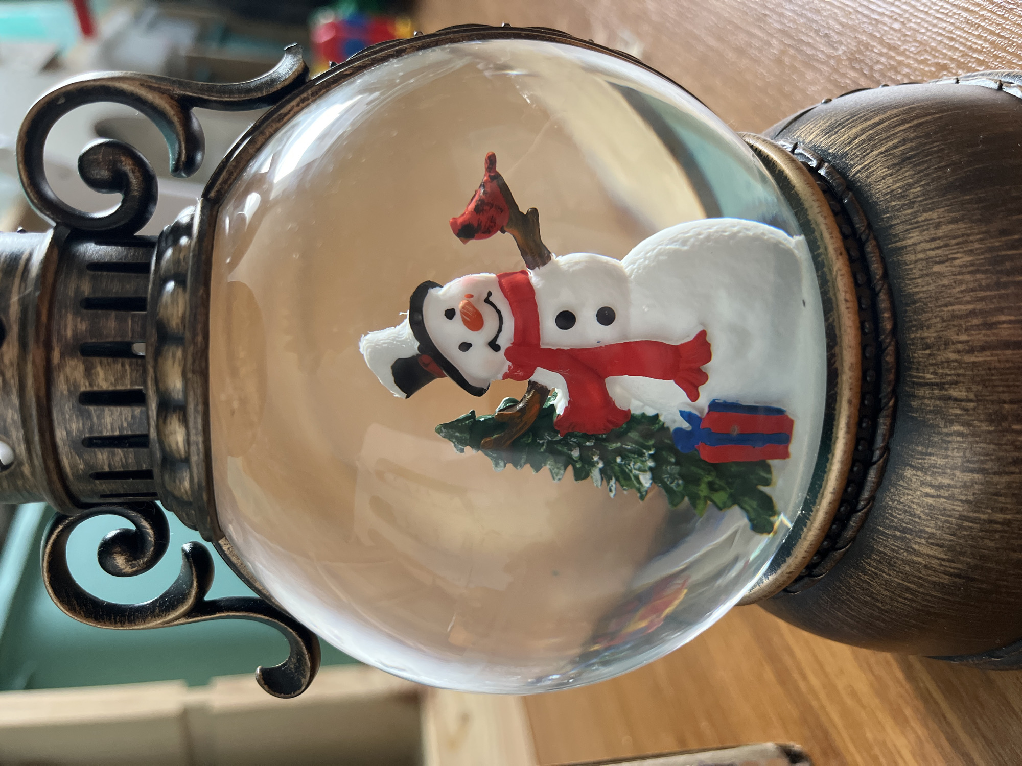The media file displays a snow globe, captured with a 90-degree clockwise rotation, making the item appear on its side. Inside the clear glass globe, a cheerful snowman is the central figure, wearing a red scarf, black top hat, and featuring a carrot nose and coal-like eyes and smile. Its cheeks are lightly flushed pink. A small red cardinal perches on a branch extending from behind the snowman's head. At the base of the snowman, a small green evergreen tree is visible alongside two wrapped gift boxes, one predominantly red and the other blue.
The snow globe is encased in an ornate, antique-bronze or dark metallic-colored base with a brushed or distressed finish. This base features intricate scrolled decorative elements on its sides, adding to its classic aesthetic. The rim where the glass globe meets the base has a detailed, possibly braided or studded, pattern.
The scene depicts a festive, winter-themed decoration. No specific activity is taking place as it is a still object. The background, visible through and around the globe, suggests an indoor setting, likely on a wooden surface, with soft light reflections on the glass. There is no visible text on the snow globe or in the surrounding area. This item is located in Chaikovsky, Russia.