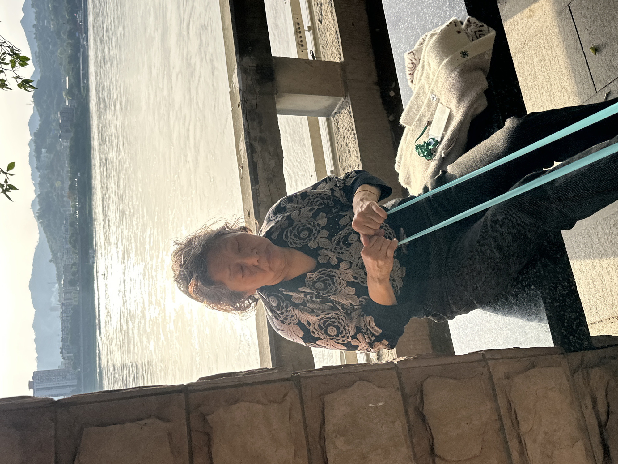 A waist-up shot shows an older woman with graying hair, dressed in a dark, floral-patterned top and dark pants, standing on a stone ledge. She is holding a light-blue resistance band with both hands. To her right, on the ledge, there is a light-colored sweater with a tag. Behind her, through concrete pillars, is a sunlit body of water, with mountains and buildings visible on the opposite shore. The scene is set in Jiangjin, China.