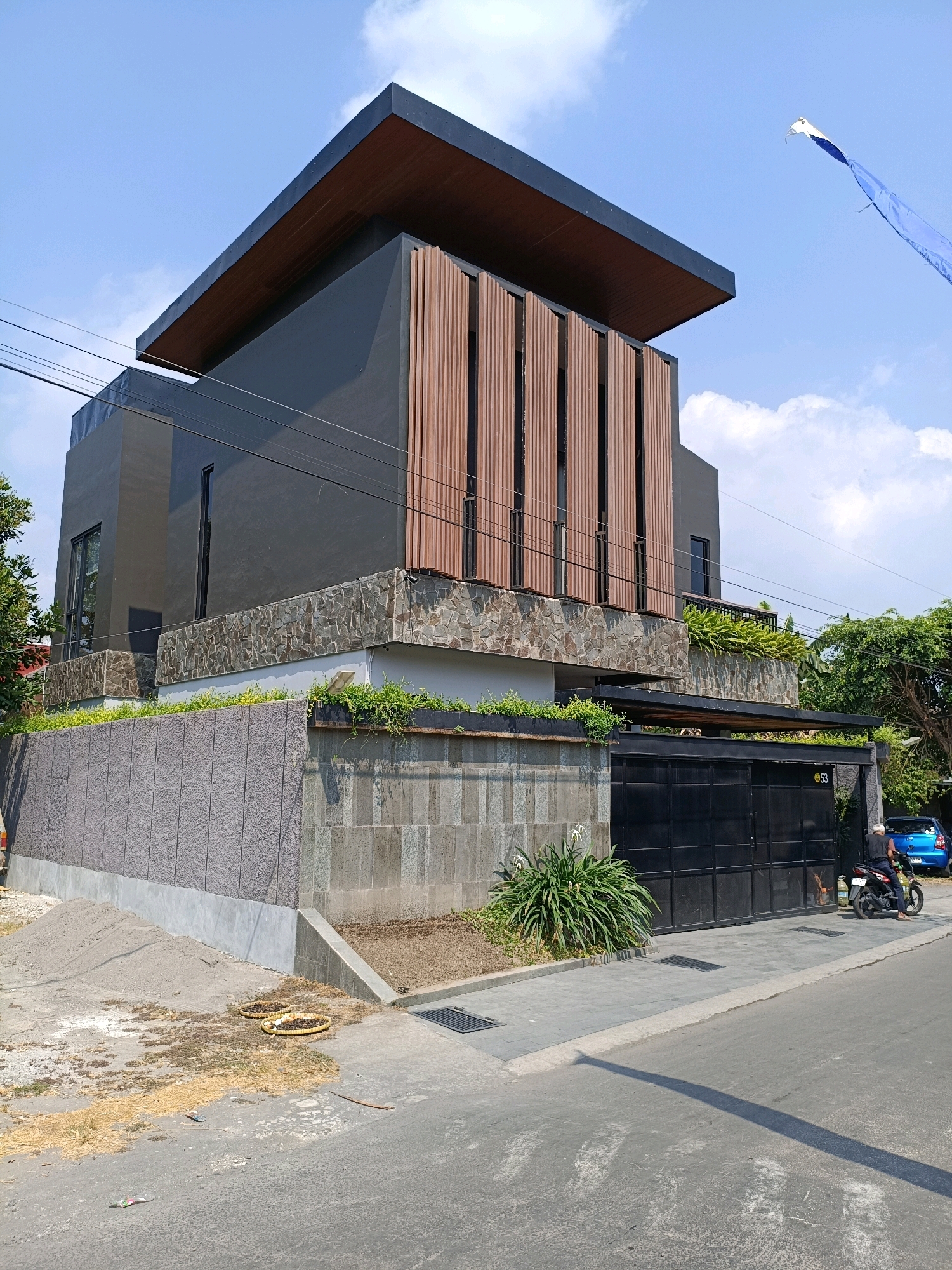 The image depicts a modern two-story house with a dark gray facade, a distinctive sloped brown roof, and vertical wooden slats on the second story. The ground floor features a stone-clad lower section and a black metal gate with the number "53" visible. A person is seated on a black motorcycle parked to the right of the gate, with a blue car parked behind them. The house is situated on a street with a gray paved road and sidewalk, with a small pile of sand and some dry vegetation to the left of the property entrance. Green plants and trees surround the house. The scene is set under a clear blue sky with scattered clouds, indicative of a daytime exterior shot in a residential area. The architectural style suggests a contemporary urban or suburban dwelling in Purwomartani, Indonesia.