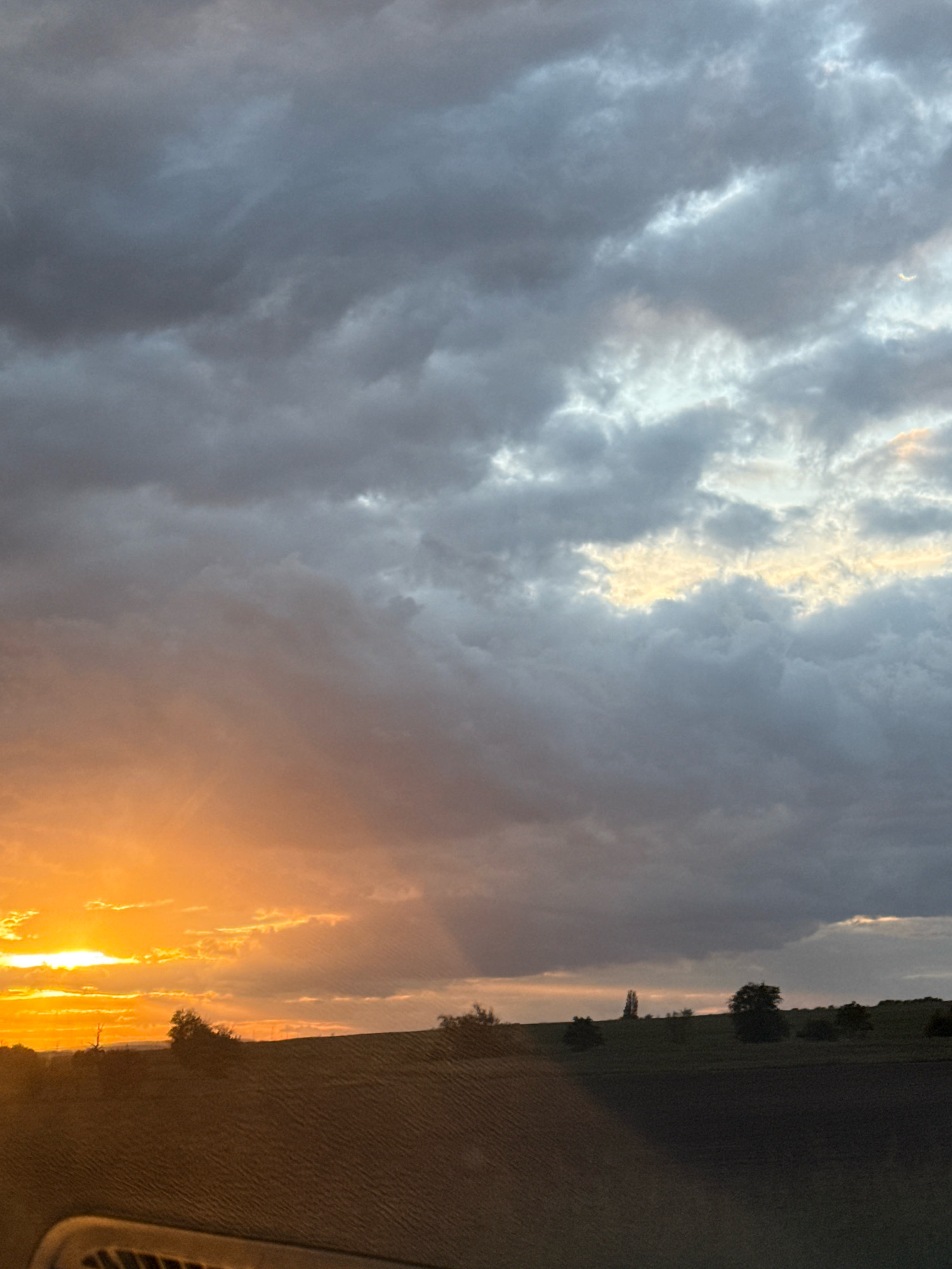 The image depicts a dramatic sunset over a rural landscape, likely in or near Clingen, Germany. The sky is filled with a mix of dark, ominous storm clouds and bright, fiery oranges and yellows from the setting sun. The sun itself is a brilliant orb on the left side of the frame, casting a warm glow on the clouds and horizon.
Below the sky, a rolling landscape is visible. The foreground is dominated by a dark, plowed field. Further back, a gentle slope rises, dotted with scattered trees and bushes that appear as dark silhouettes against the sky. The overall impression is one of natural beauty and power, with the forces of nature on full display. The scene is devoid of any people or human activity, emphasizing the vastness and solitude of the natural environment.