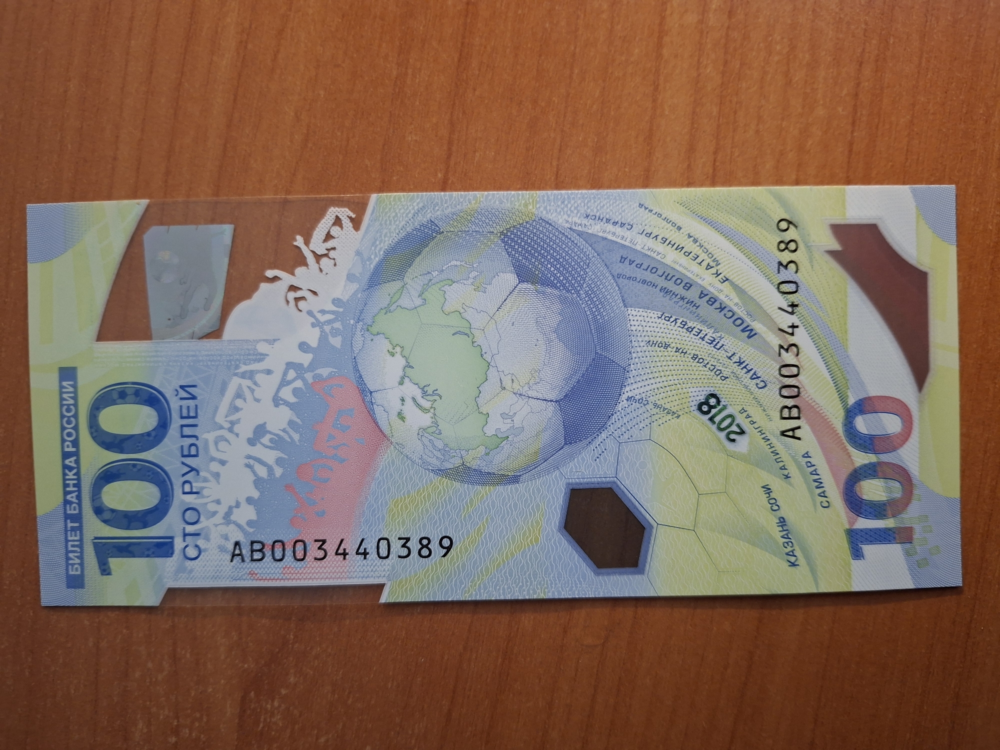 A Russian 100 ruble banknote is pictured lying on a wooden table. The banknote has a light blue and yellow design featuring a globe shaped like a soccer ball, outlines of cheering crowds, and text in Russian. The denomination "100" is prominently displayed in large blue numbers at the bottom and top of the bill. A serial number, "AB003440389," is visible vertically on the left side of the banknote. The year "2018" is also visible on the banknote. The background is a plain wooden surface with visible grain.