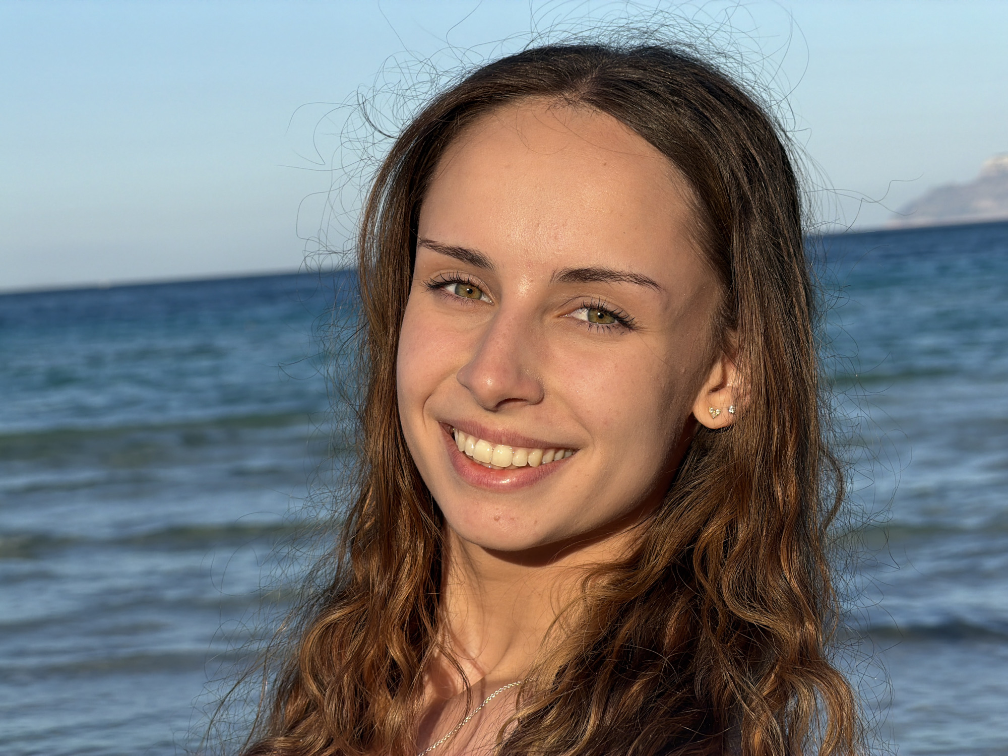 A young woman with light brown hair and green eyes stands smiling against a background of a blue sea and sky. She has a fair complexion and is wearing stud earrings. The location appears to be a beach, possibly near Alcúdia, Spain, given the coastal environment. There's a hint of land visible in the far distance.