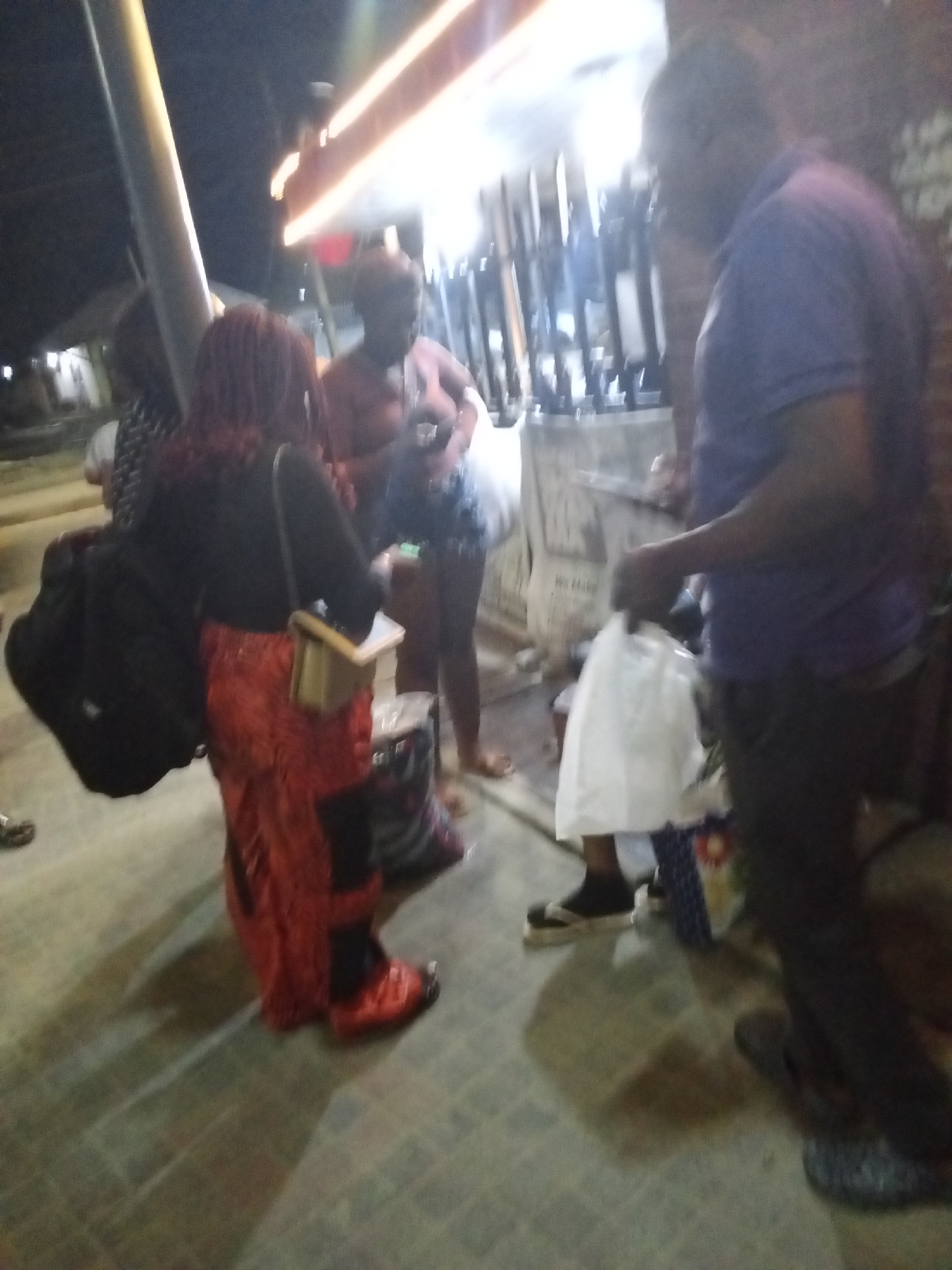 The scene is located in Ikorodu, Nigeria, in an outdoor public area at night. A group of people are gathered near a brightly lit structure, possibly a shop or kiosk.
A man in a purple shirt and dark pants is standing to the right, holding a white plastic bag. He seems to be interacting with the kiosk. To his left, a woman with short hair is wearing denim shorts and a light colored top and standing near a shopping bag. Another woman with red hair is facing away, wearing a long colorful skirt, a black shirt, and carrying a backpack. She is also holding a small box.
The setting is likely a street or roadside, with a metallic support pole visible. The ground appears to be paved.