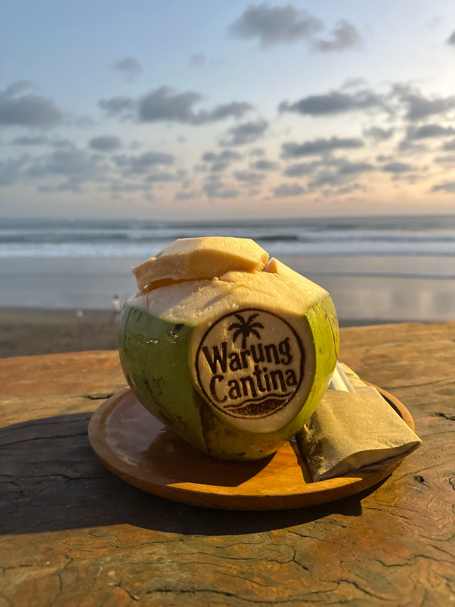 A freshly opened young coconut with a carved logo featuring a palm tree and the text "Warung Cantina" rests on a wooden plate. The coconut, filled with coconut water and topped with slices of coconut flesh, is placed on a rustic wooden table. In the background, the ocean meets the sky under a cloudy sunset. The scene is set on a beach in North Kuta, Indonesia. A small brown paper packet sits beside the coconut on the plate.