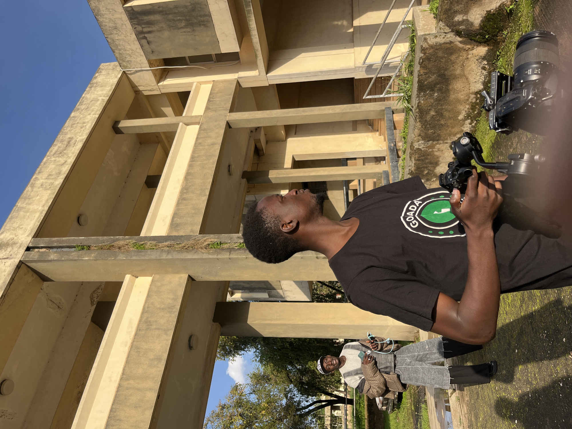In a shot with a vertical orientation, a young Black man, wearing a black t-shirt with a circular green logo and white text, looks upwards. He is holding a camera, which is attached to a gimbal. The background features a large, Brutalist-style concrete building with visible beams and supports, suggesting an outdoor setting under a clear blue sky. Sunlight casts shadows, indicating it's daytime, likely morning or afternoon. To the right of the main subject, another person is partially visible, sitting and holding a phone. The overall scene appears to be in an urban or semi-urban environment, possibly an institutional or educational complex, given the architecture. The individual's upward gaze and the presence of camera equipment suggest they might be filming or directing a shot. The concrete structure has patches of greenery growing on ledges, hinting at neglect or natural overgrowth.