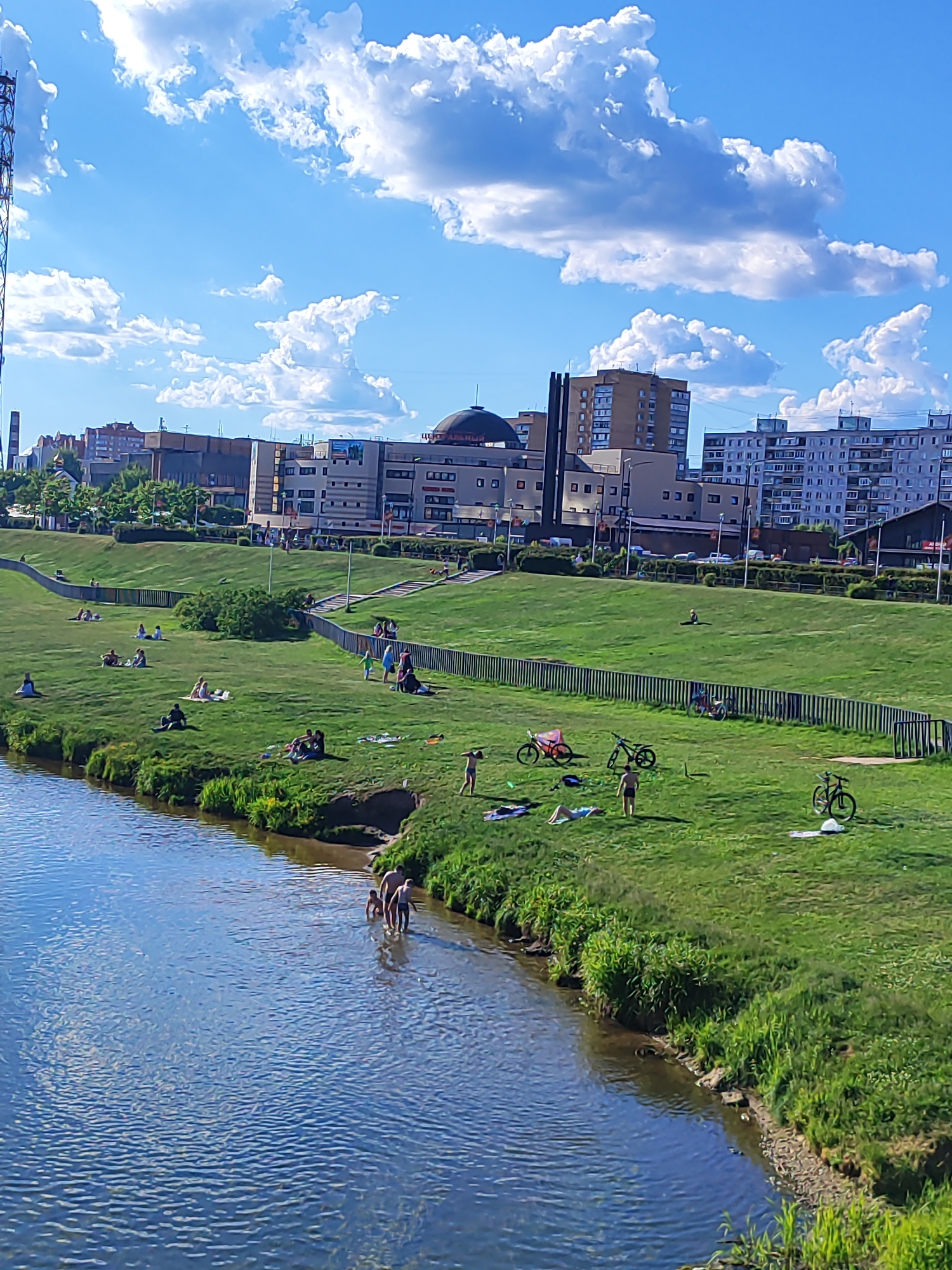 This media file depicts a vibrant outdoor scene on a sunny day, showcasing people enjoying a riverside park in an urban setting, likely Shchyolkovo, Russia. The image is taken from an elevated viewpoint, looking down upon a calm river and its green, sloping bank, with city buildings forming the backdrop.
The sky is a brilliant blue, dotted with large, fluffy white cumulus clouds, indicating clear and pleasant weather conditions, likely during daytime. On the right, a wide, well-maintained grassy embankment slopes down to the river's edge. Numerous individuals and small groups are scattered across the grass, engaging in various leisurely activities. Many are seen relaxing, sitting, or lying on blankets or directly on the grass, suggesting sunbathing or picnicking. A few people are walking on the upper parts of the bank.
A black metal fence runs horizontally across the middle ground, separating the lower grassy area from a higher section behind it, where more parkland or urban infrastructure is visible. In the background, a cluster of multi-story buildings, including residential blocks and commercial structures, defines the urban landscape. One notable building features a distinct dark dome-shaped roof. Partially visible Cyrillic text, possibly "ЦЕНТР" (Tsentr, meaning Center), can be seen on one of the buildings. To the far left, a tall metal lattice tower rises against the sky.
Closer to the river, several bicycles are parked on the grass, indicating a mode of transport for some visitors. At the immediate water's edge, a small group of people, primarily men, are wading or swimming in the river's clear, shallow waters, adding to the recreational atmosphere. The overall scene conveys a sense of relaxation and community enjoyment of nature amidst an urban environment. The lush green grass and calm river contribute to a serene and enjoyable setting.