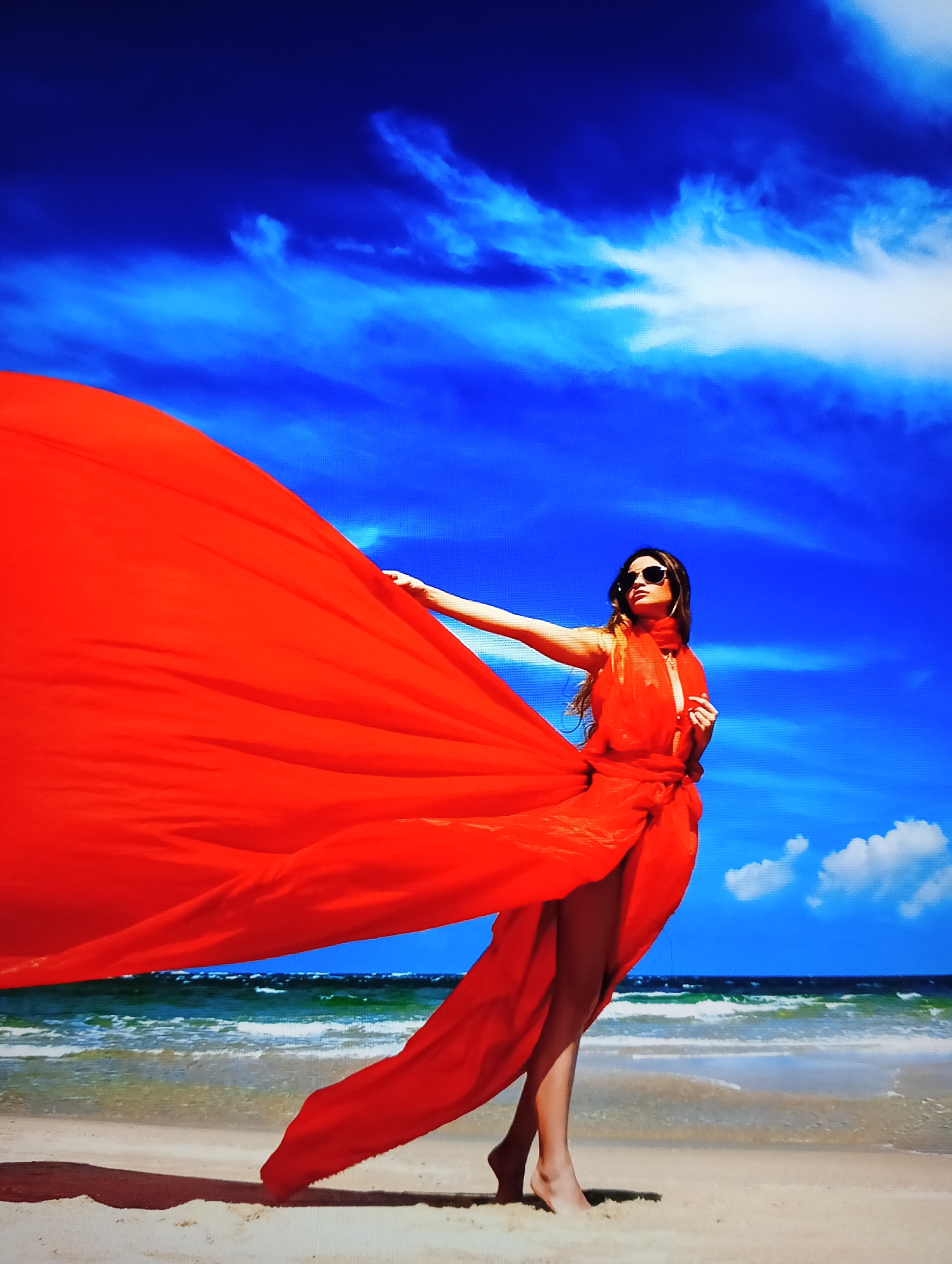 The image shows a woman standing on a beach. She is wearing sunglasses and a red dress that is billowing in the wind. The background includes a blue sky with some clouds and ocean water. The location is Nha Trang Ward, Vietnam, as evidenced by the typical beach environment.