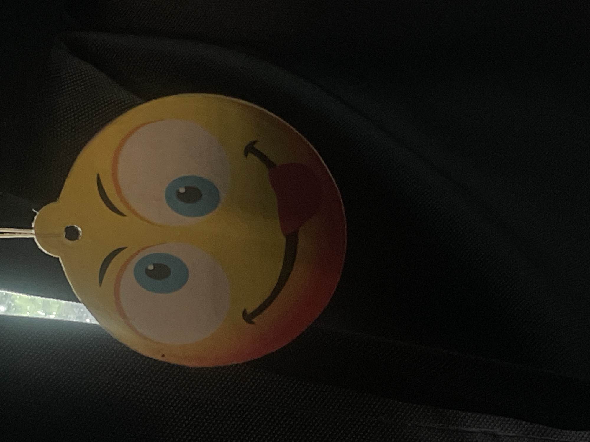 The image shows a close-up of a yellow smiley face air freshener hanging from a string. The air freshener has large blue eyes with white pupils and a wide mouth with its red tongue sticking out. The smiley face also has rosy cheeks. The background is dark and appears to be fabric, possibly from a car's interior. There is a vertical light source visible in the upper left corner of the image. There is no information in the image to indicate its location in Ganderkesee, Germany.