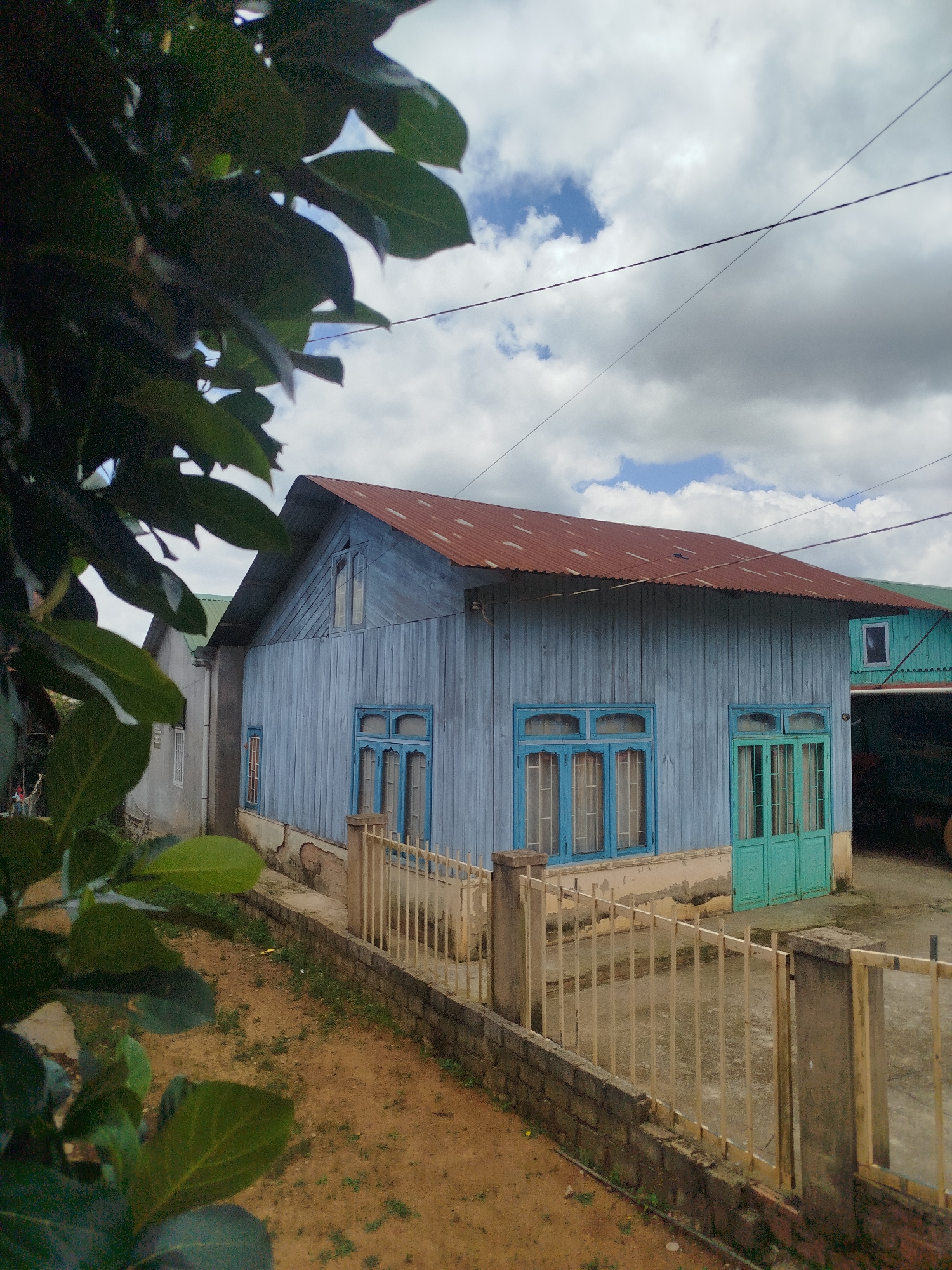 A light blue wooden house with a rusty corrugated metal roof is visible in Thôn 5, Vietnam. The house has several blue-framed windows and two teal-colored doors. A white metal fence with brick posts runs along the front of the property, bordering a dirt path with sparse vegetation. The sky is cloudy with patches of blue visible. Dark green leaves from a tree are in the foreground, partially obscuring the view of the house. To the left of the blue house is a lighter-colored building with a green roof.