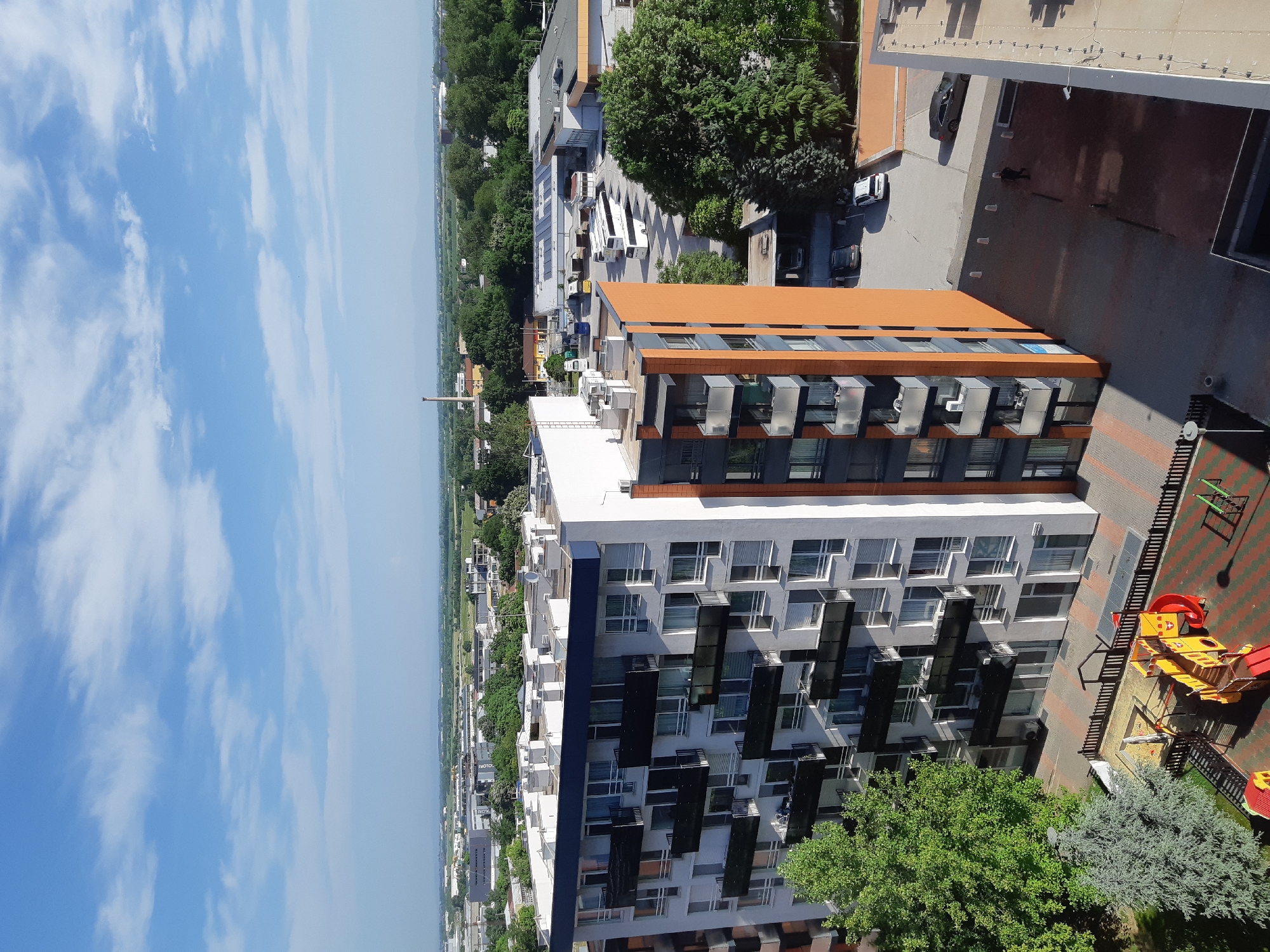 The image shows an aerial view of a modern apartment building in Plovdiv, Bulgaria. The building has a distinctive design with alternating sections of white, grey, and orange facade, and numerous balconies with air conditioning units.
Adjacent to the building is a children's playground with red and yellow equipment. To the left of the building, there is a clear view of the city's skyline and surrounding landscape under a partly cloudy sky. Other buildings and green spaces can be seen in the background. Several cars are parked nearby, suggesting a residential area.