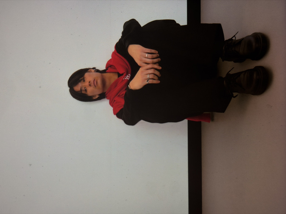 The image depicts a single individual, male, seated on a light grey floor and leaning against a light off-white wall. The individual, appearing to be of East Asian ethnicity, possesses dark, medium-length hair and is looking directly forward. They are attired in a black long-sleeved top, black wide-leg trousers, and black lace-up shoes. A red scarf, displaying white markings or insignia, is draped over their shoulders. Multiple rings are visible on both hands, and small earrings are present. The wall features a thin, dark horizontal line near the bottom, marking the transition to the floor. The setting is an indoor, minimalist environment with even illumination. There are no discernible architectural features or external elements to identify a specific location within London, United Kingdom.