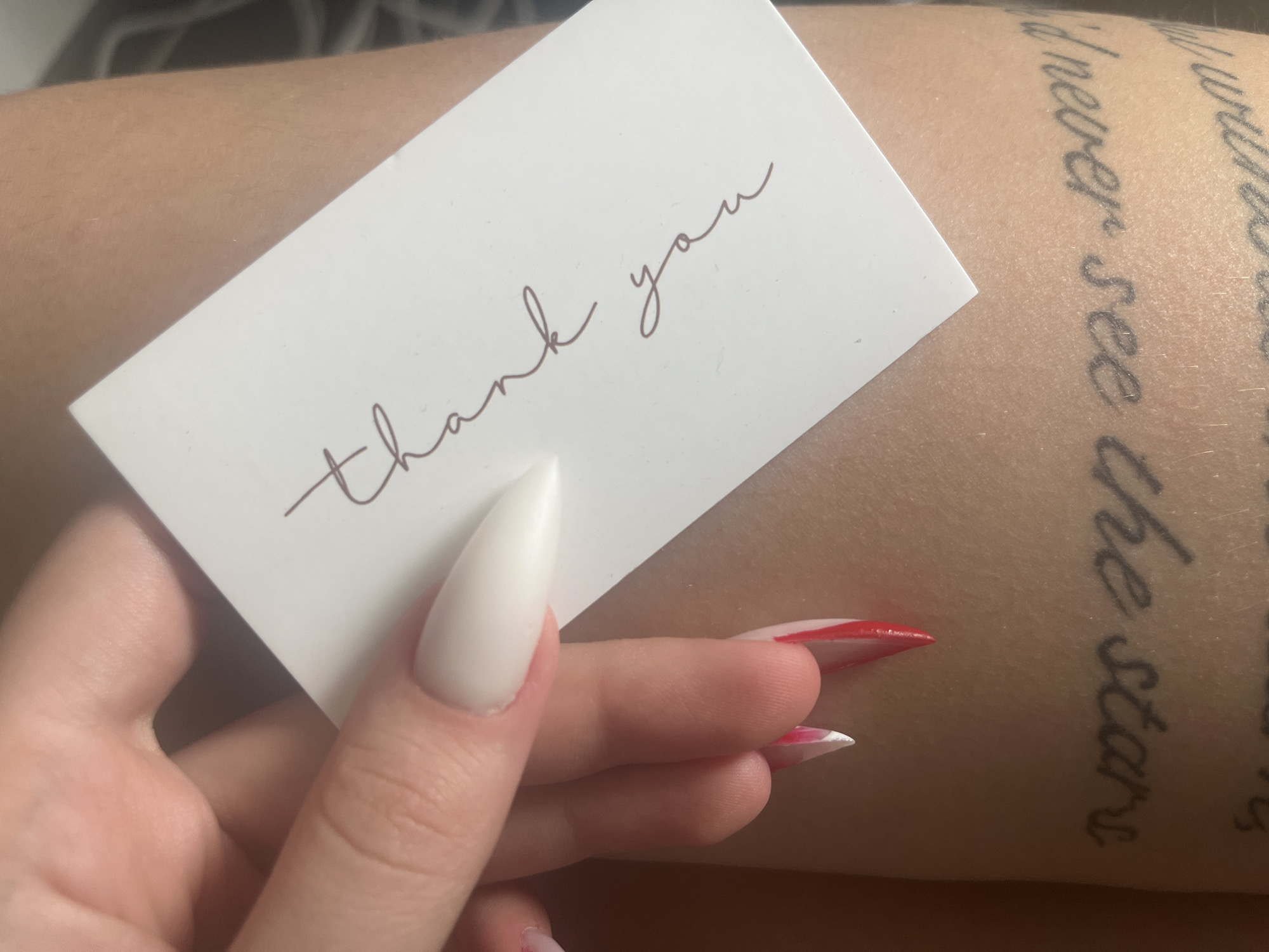 A pale-skinned arm with a tattoo of text is shown in a close-up shot. The tattoo is partially visible with the words "never see the stars" appearing legible. A white business card with the words "thank you" written in a cursive font lies on the arm and is being held by two fingers of the same hand. The nails on the fingers are long and manicured; one nail is white, and the other has a red and white design. The background is out of focus, and the location is not discernible from the image. The location of Varna, Bulgaria, cannot be verified from the provided image.